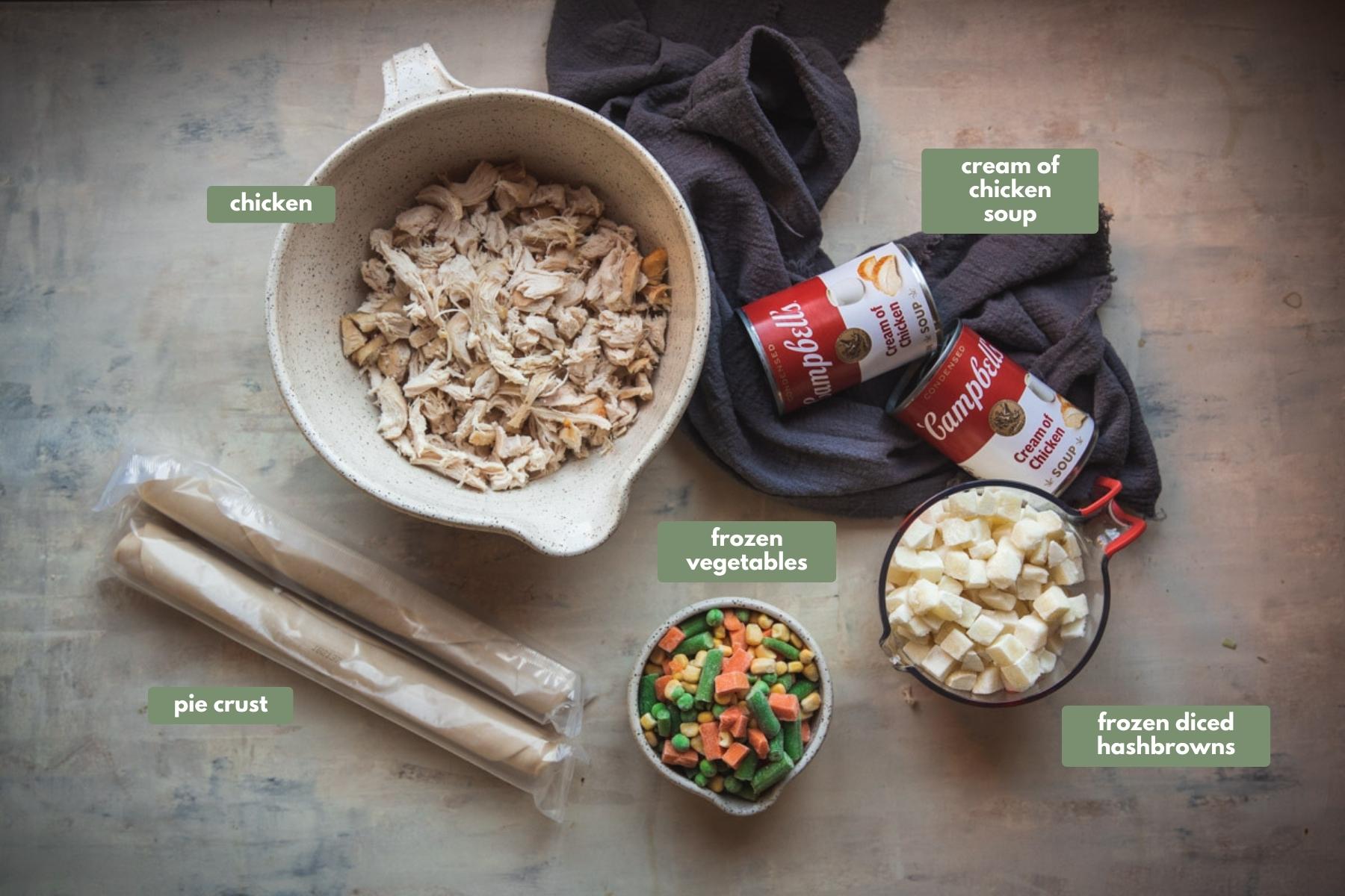 All the ingredients required to make this recipe sitting on a grey counter. There is a large white bowl containing cooked chicken, 2 tins of campbells cream of chicken soup, a glass jug filled with frozen hash brown chunks, a white bowl stuffed to the top with frozen veggies like carrot, green beans and corn. There is also 2 sheets of rolled up pie crust.