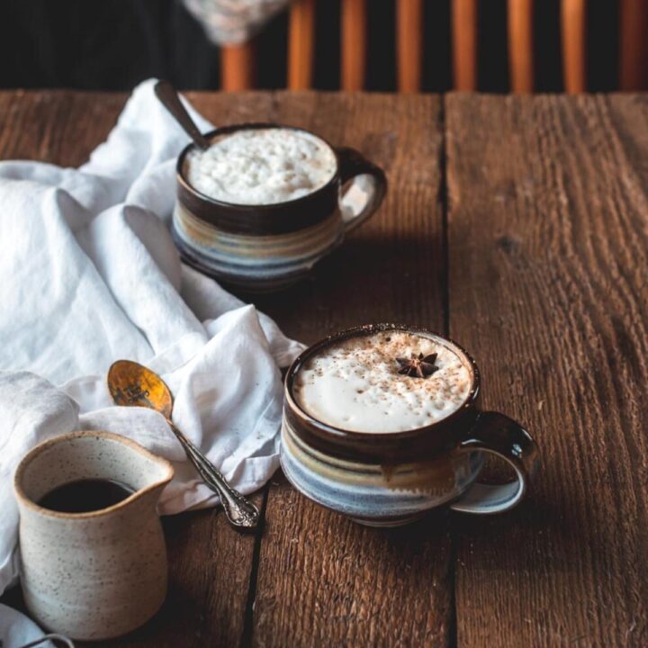 Two coffee mugs filled with foamy milk and sprinkled with cinnamon on top of a rustic wood surface. A ceramic carafe and antique spoon are next to the mugs on a white linen napkin. A wooden chair draped with a scarf is in the background.