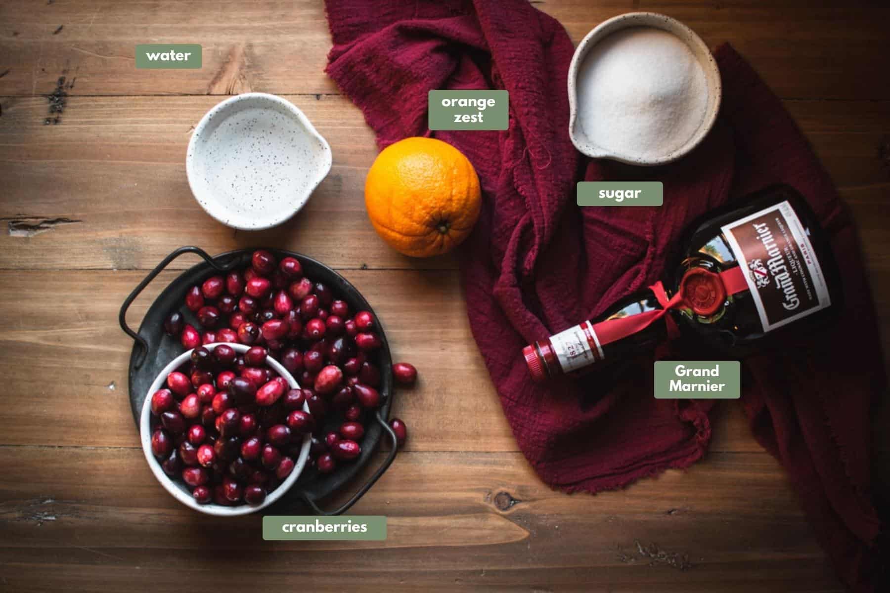 The ingredients required for this recipe lying on a wooden table. There is a whie terrazzo bowl with water inside and a black cast iron bowl filled with cranberries. A whole orange lies next to some red fabric draped over the corner of the table where a bottle of Grand Marnier sits next to a bowl of white sugar. 