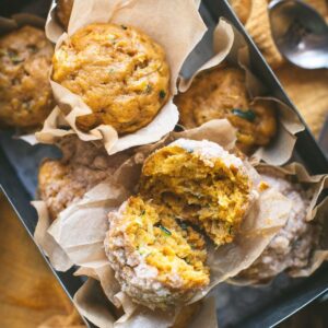 Pumpkin and zucchini muffins in parchment paper wrappers stacked inside a rectangular tin tray over a yellow linen napkin. One of the muffins is split in half to show the texture of the muffin.