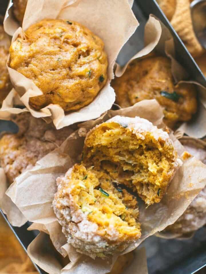 Pumpkin and zucchini muffins in parchment paper wrappers stacked inside a rectangular tin tray over a yellow linen napkin. One of the muffins is split in half to show the texture of the muffin.