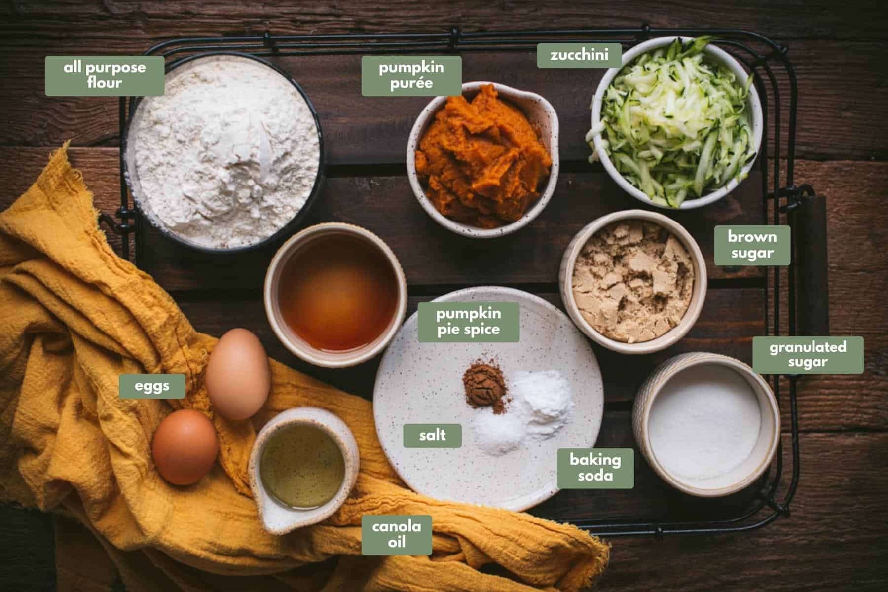 Labeled ingredients in an overhead photo next to a yellow linen napkin and placed on a wooden board.  Flour, pumpkin puree, grated zucchini, brown sugar, white sugar, vanilla, spiced, baking soda, oil, and eggs.