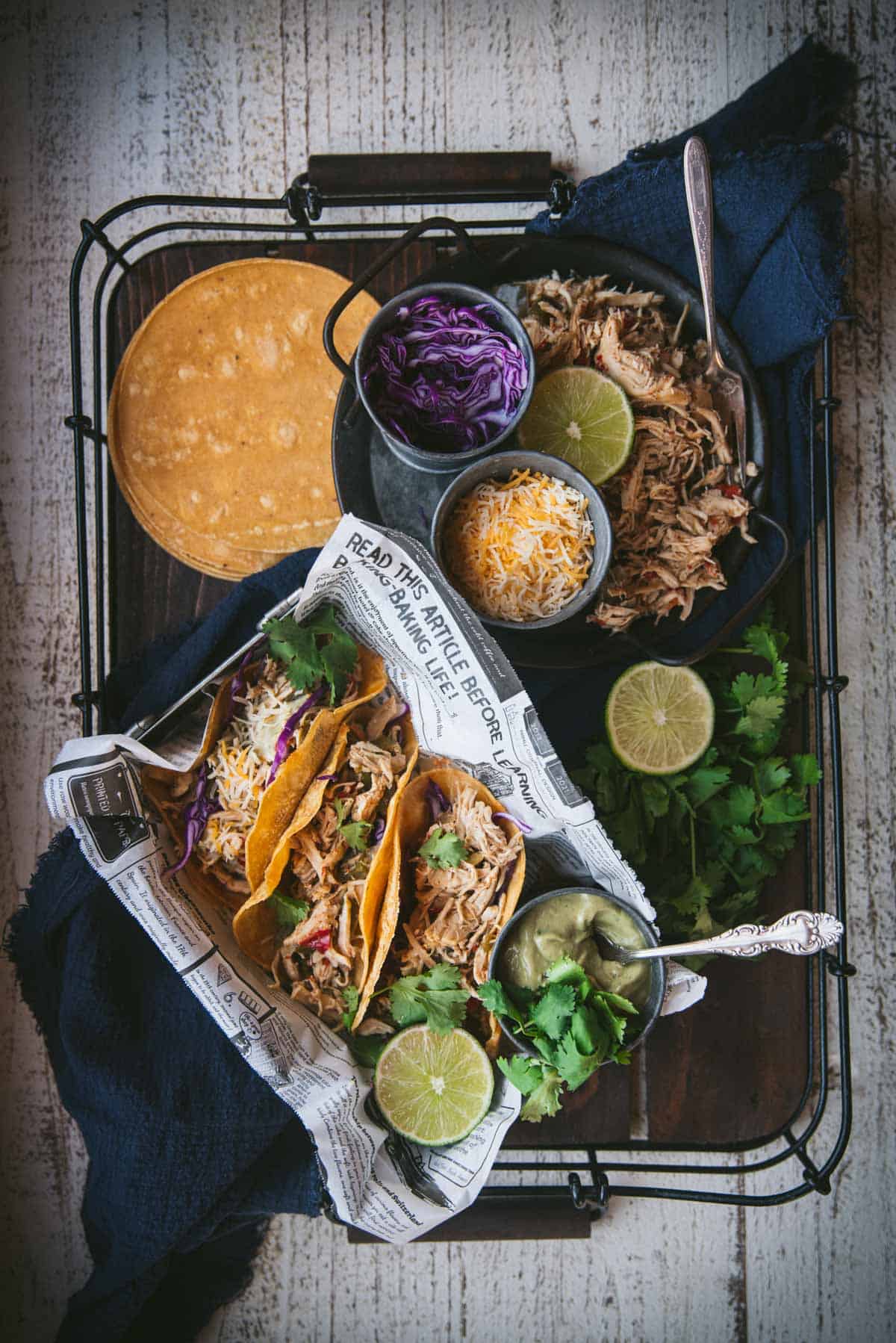 A large wooden tray with black metal handles is laid out with all the tacos and toppings. There is a newspaper lined box containing 3 chicken tacos topped with red cabbage, cheese and cilantro. Inside the box, next to the tacos is a a lime half and a small black ramekin dish filled with guacamole with a silver spoon in it. On the rest of the tray there is a black plate with more crock pot chicken on top, a lime ring, a black bowl of red cabbage and a dark colored bowl filled with cheese. Left over taco shells are piled on top of each other in the corner of the tray and extra cilantro and lime is scattered over the rest of the tray.