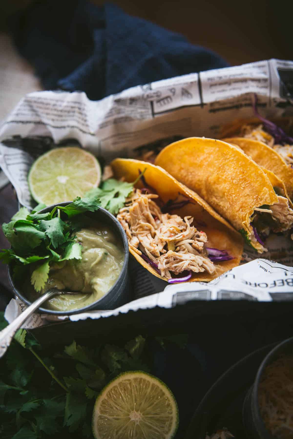 Close up image of newspaper lined box filled with 3 tacos, you can only just see the third taco in the background. The middle taco has had a bite taken out of it. Next to the tacos, nearest the camera, is a small black bowl filled with smooth guacamole and a silver spoon, there is a lime next to it and cilantro garnishing everything.