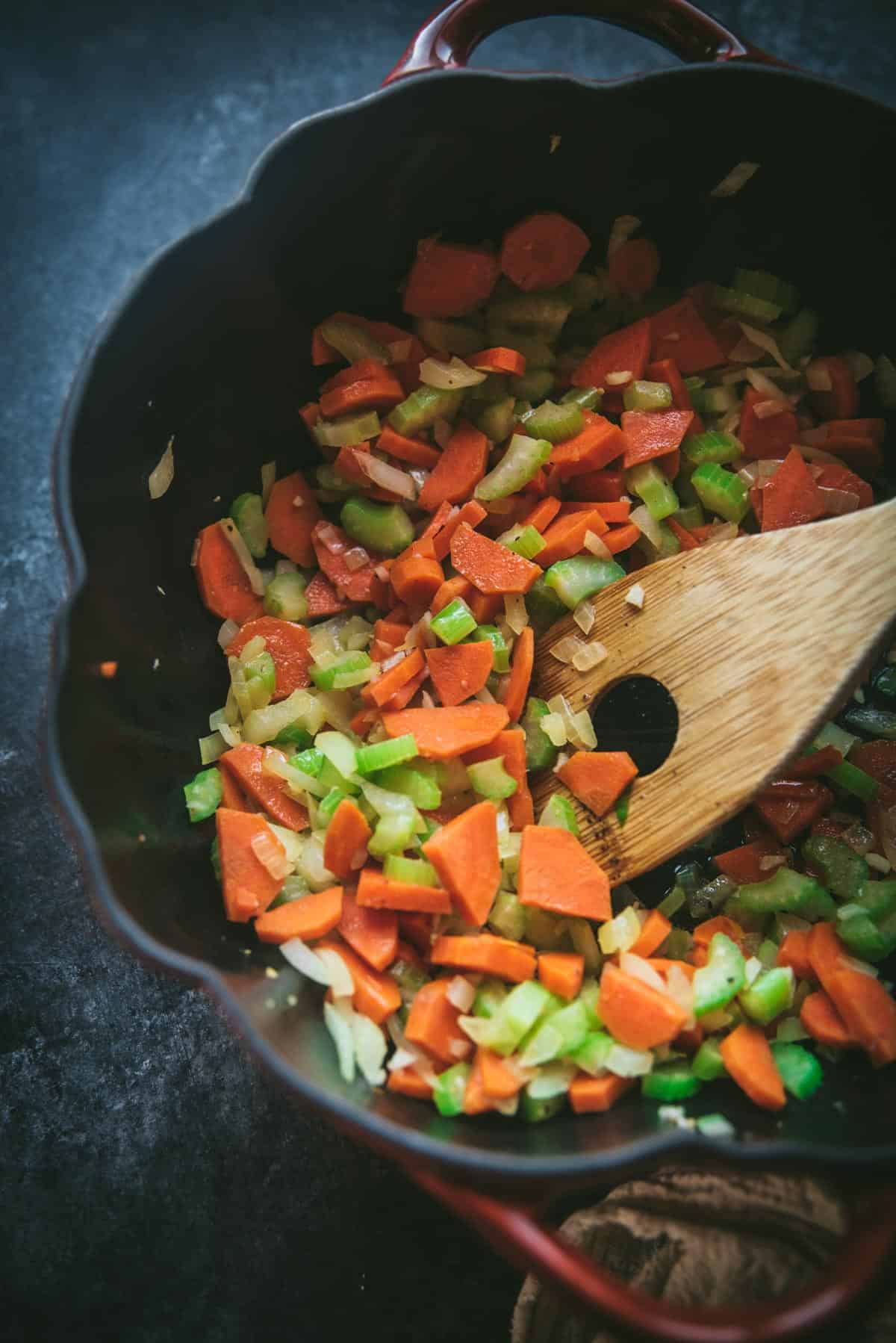 In a black skillet, carrots, celery, onion and garlic are being stirred with a wooden spoon.