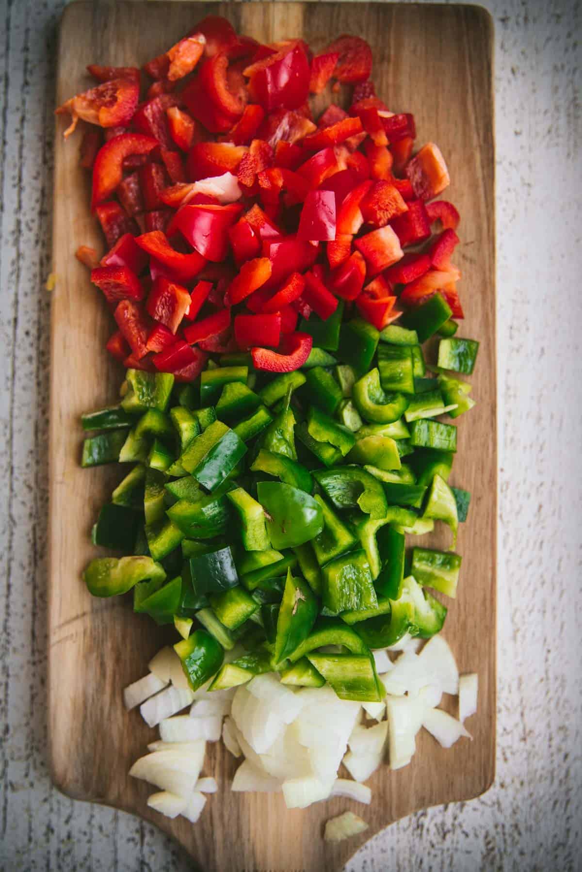 On a wooden chopping board with a handle, red pepper, green pepper and onion have been diced and are laying next to each other on the board.