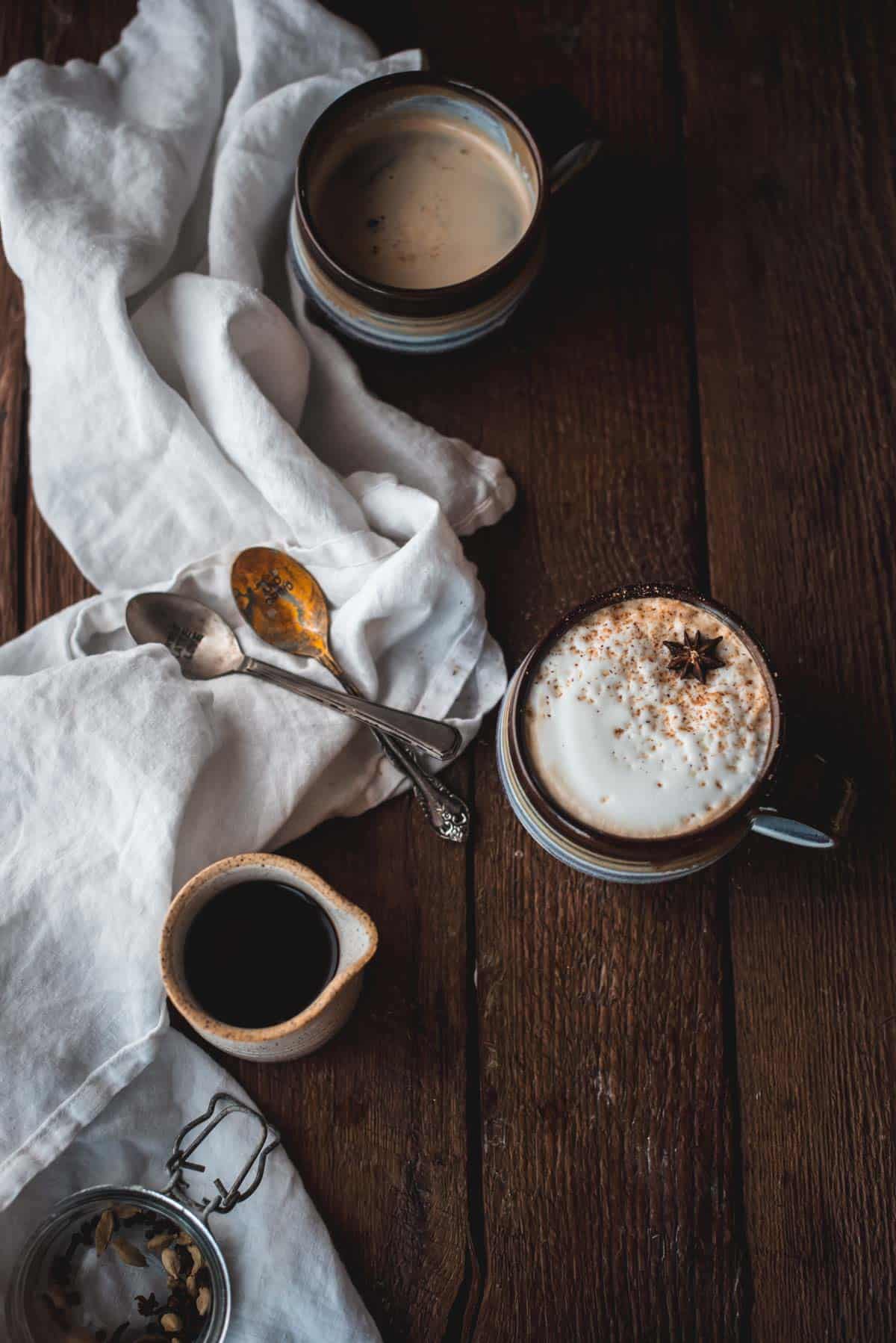 Close up overhead image of a latte in one mug and another much with chai latte and espresso but no additional milk.  The mugs are next to antique spoons on a linen napkin and a carafe filled with chai latte.  There is also a jar with chai spices.