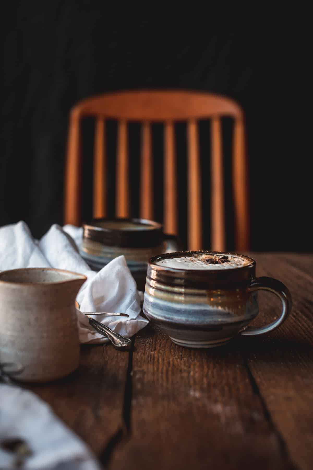 Close up image of the side of a mug filled with dirty chai latte.  The milk on top is very foamy and sprinkled with cinnamon and an anise seed pod.  Next to the mug is a linen napkin with spoons on it and a ceramic carafe.