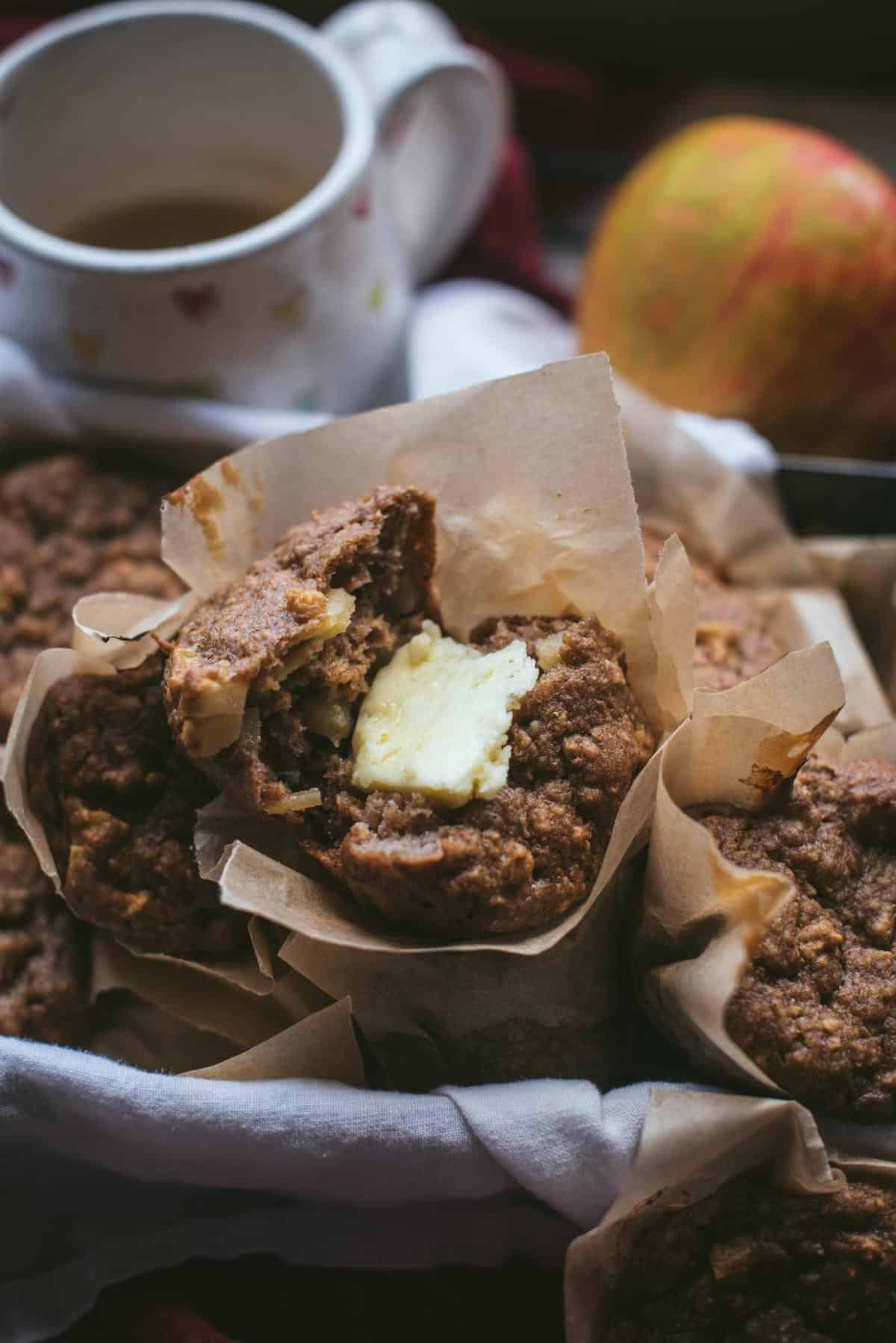 A pile of apple muffins lined in brown papers in a cloth lined container. One of the muffins has been broken in half and a chunk of butter is on top of it.