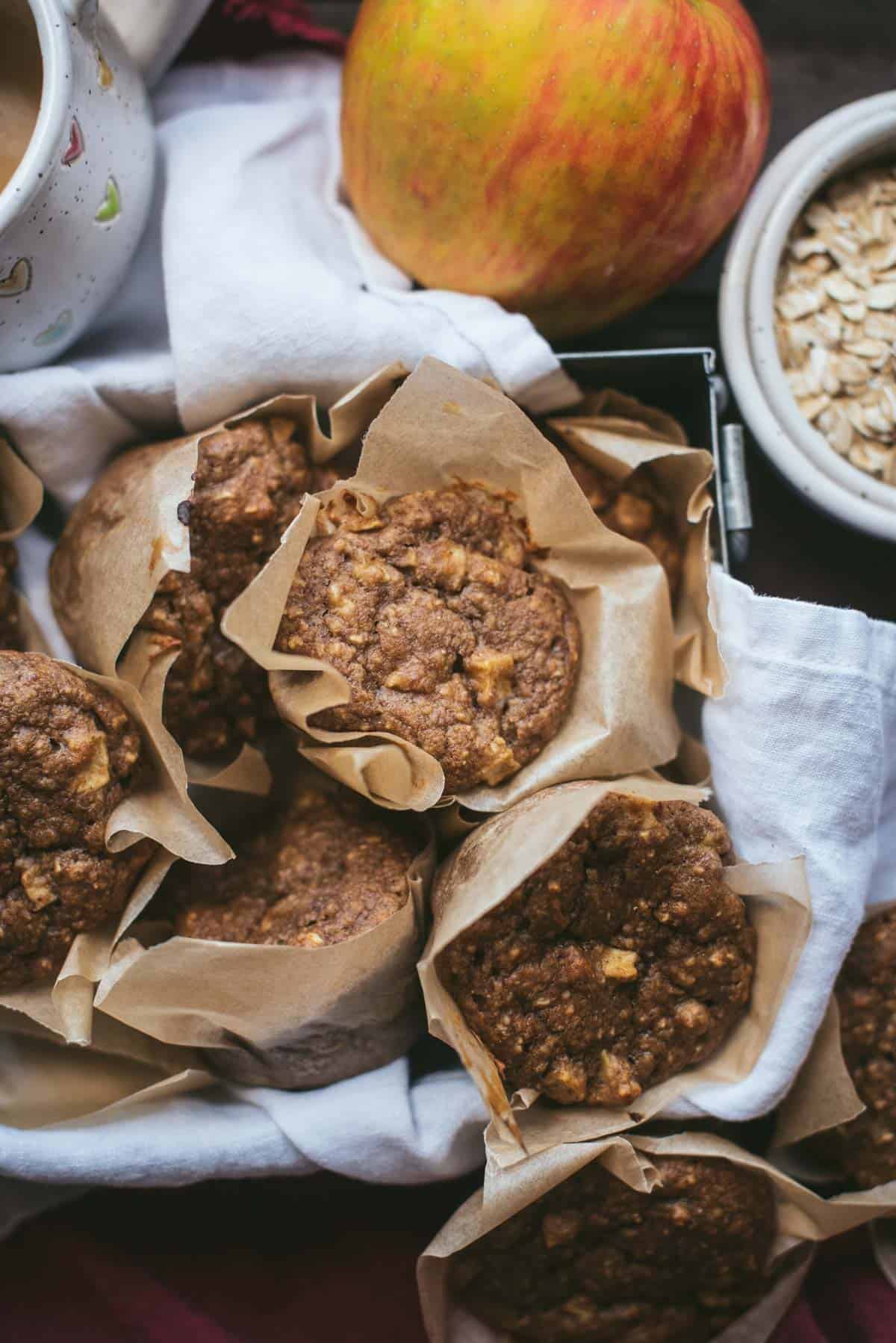 Healthy apple muffins, wrapped in brown paper sitting in a deep silver tray lined with white cloth. There is an additional apple in the corner as well as some leftover oats.