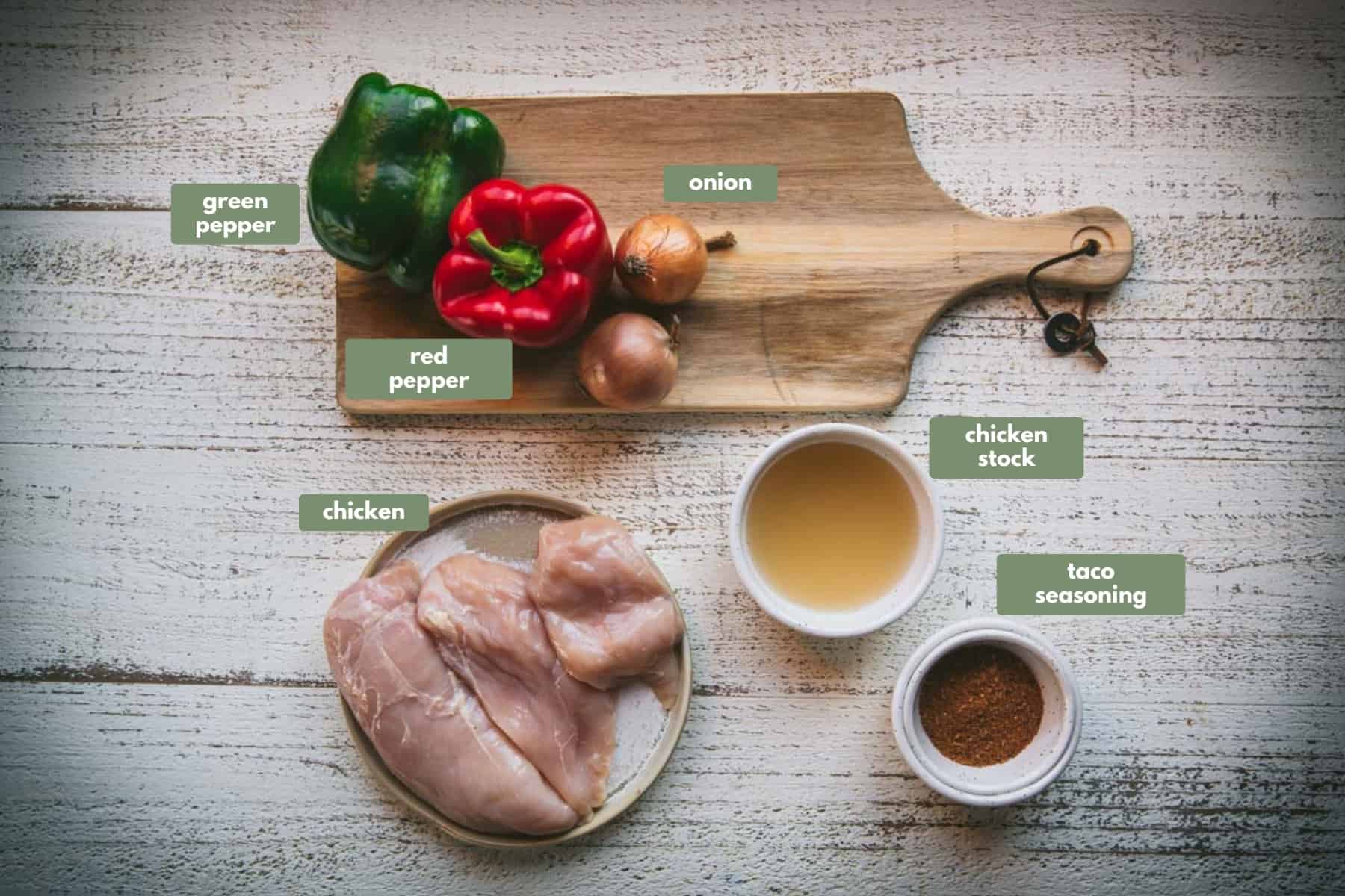 All the ingredients required to make crock pot chicken for inside your tacos. On a white wooden  table is a ceramic plate with raw chicken, a white bowl of chicken stock and a small white bowl with taco seasoning. There is a small wooden chopping board with a red and green pepper and 2 brown onions sitting on the edge of the board.