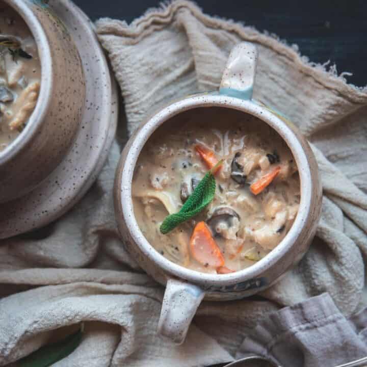 Creamy chicken and rice soup sitting in a deep cream ceramic bowl. You can see carrots and mushrooms in the soup and it is garnished with fresh herbs. In the corner you can see the edge of another bowl and they are sitting on a cream colored cloth next to 2 silver spoons.