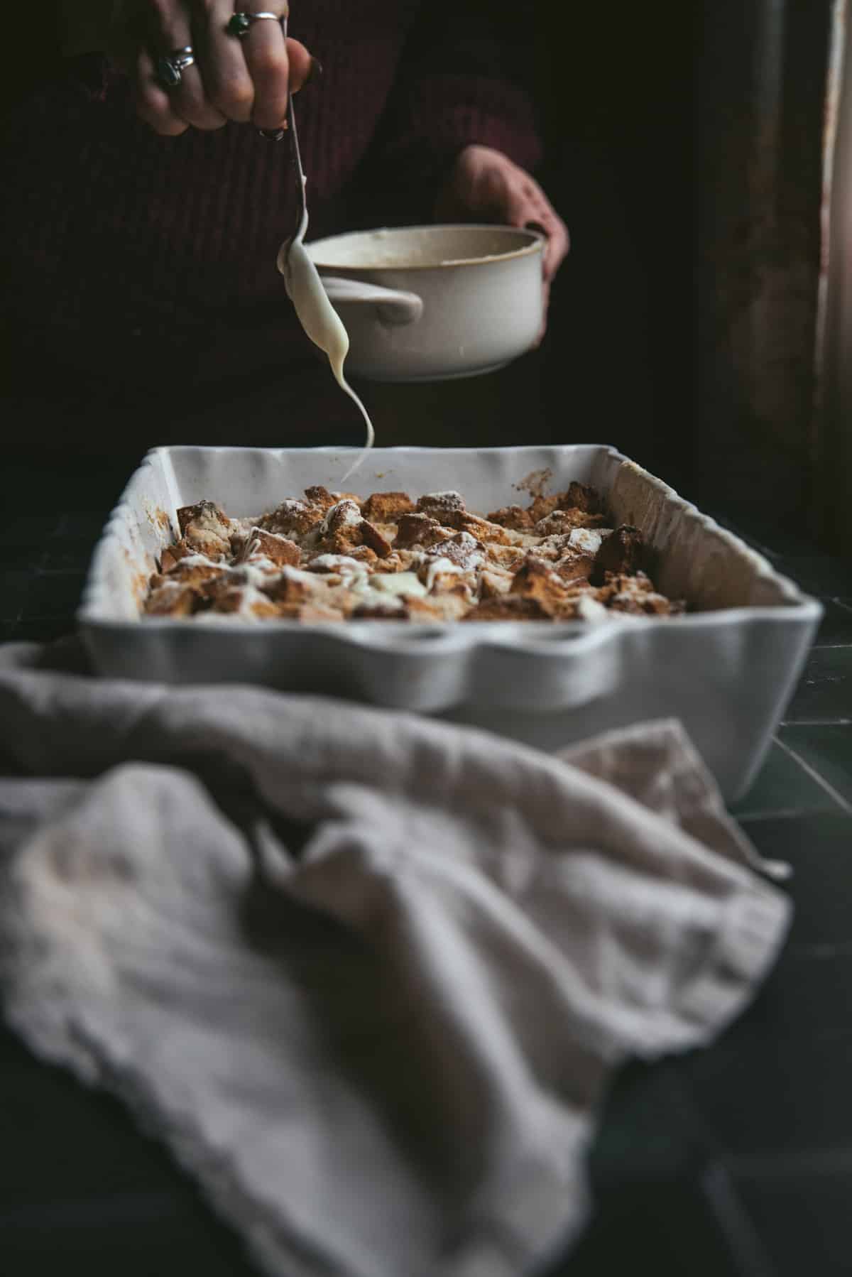 Spoon drizzling glaze over baked french toast in a white baking dish.  In the foreground is a tan linen napkin.