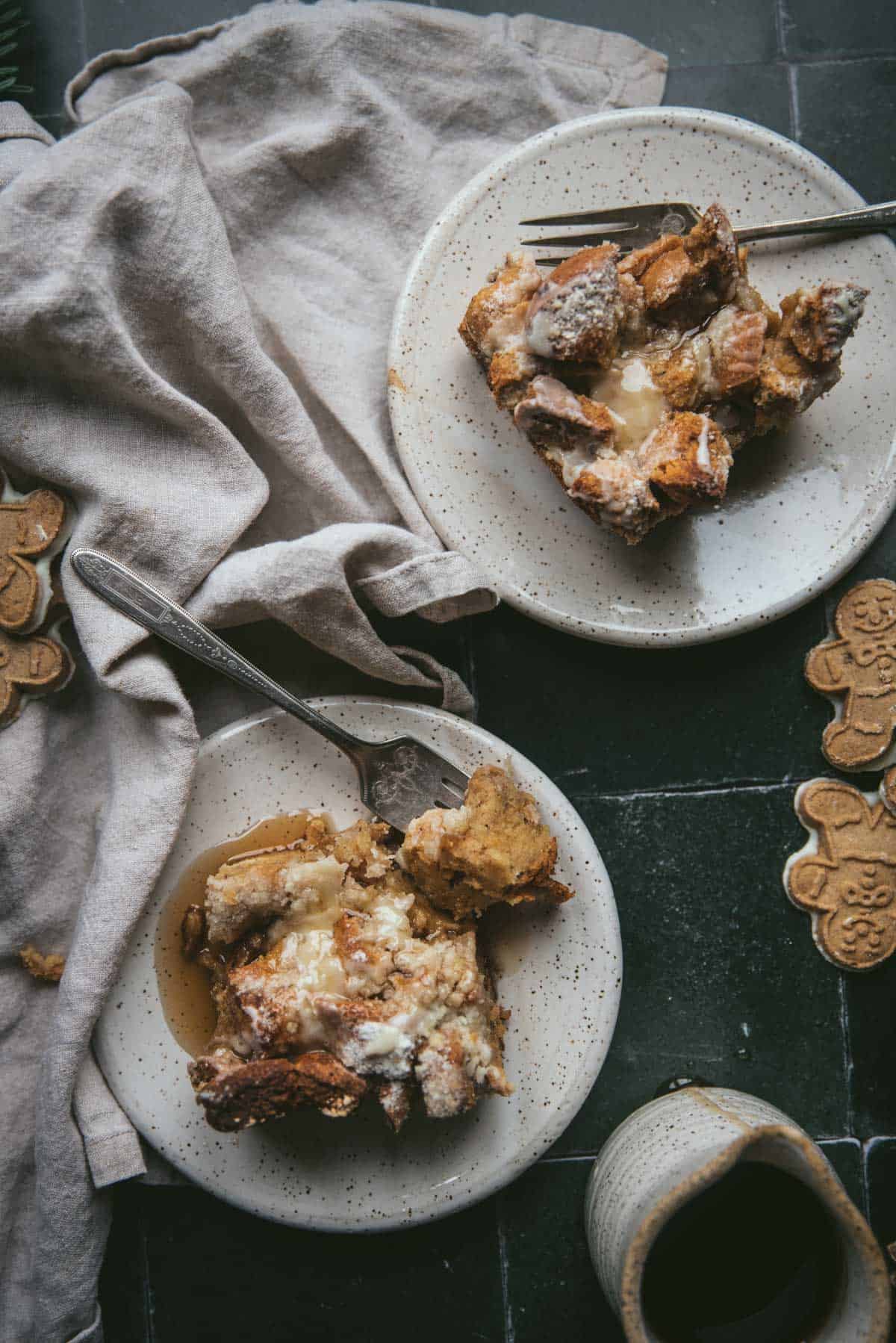 Overhead image of two white speckle ceramic plates each holding a piece of gingerbread french toast casserole drizzled with maple syrup.  Plates are on a green tile surface with linen napkins, gingerbread cookies, and a ceramic pourer next to the bottom plate.