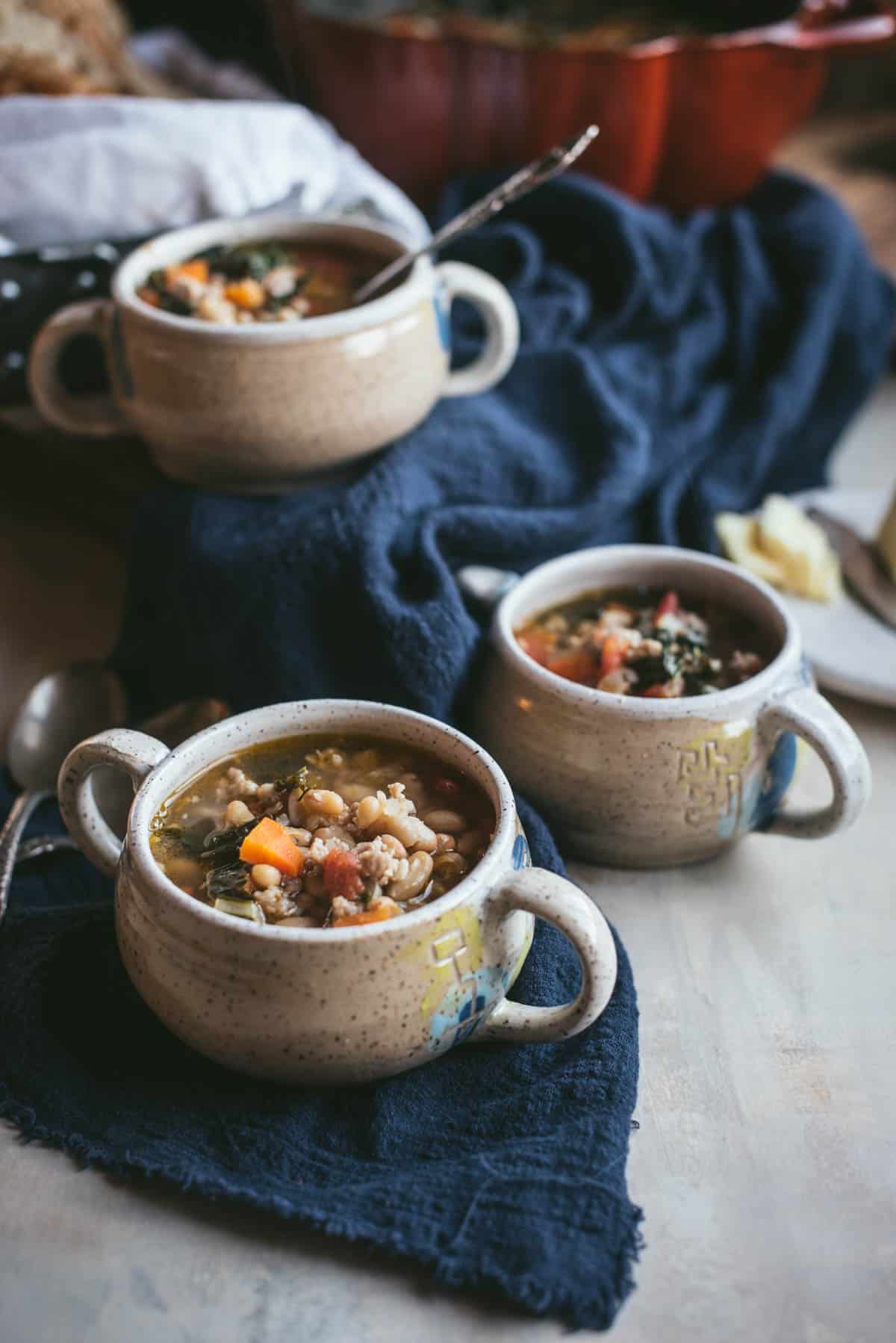 A blue dish towel is on a grey counter, on top of the cloth are white bowls with handles, they are filled with sausage bean and kale soup. There are some spoons left on the counter.