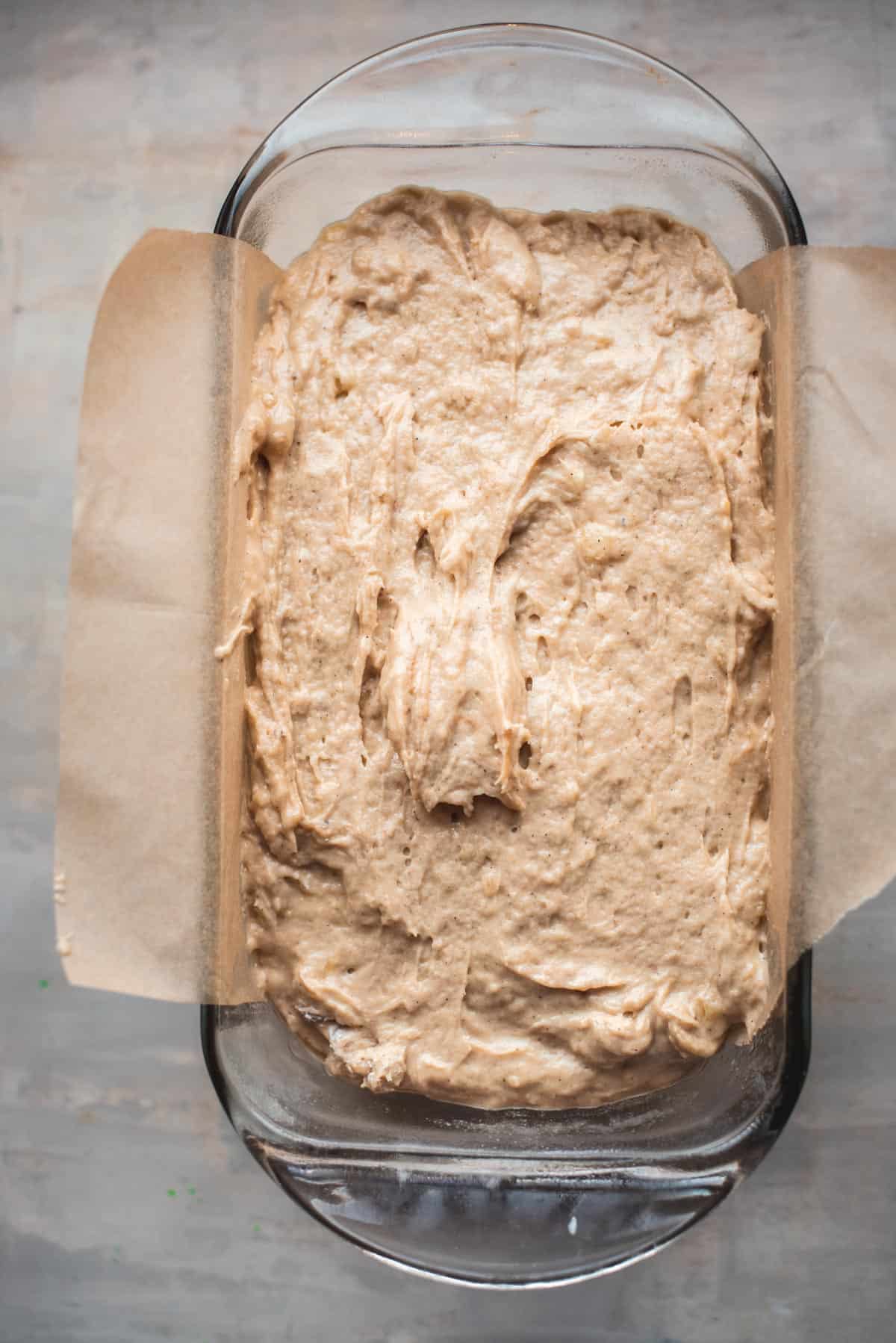 The cake batter has been placed into a parchment lined, glass loaf pan and is sitting on a grey counter.