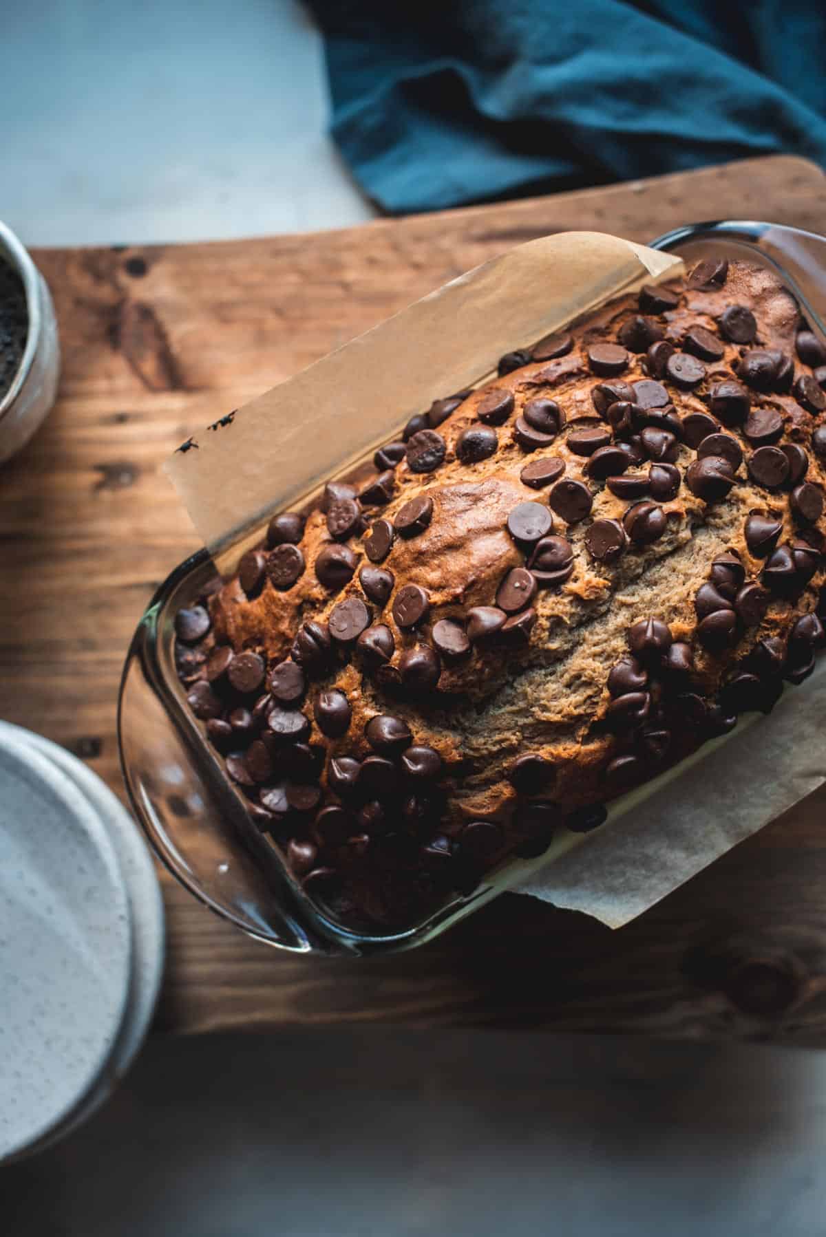 View from above. The glass loaf pan is placed onto a wooden chopping board. The Peanut butter banana bread has been baked and has risen in the dish, there are lots of chocolate chips on top and it is slightly cracked along the middle.