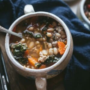 A deep white double-handled bowl is filled to the top with Sausage, White Bean, and Kale Soup. You can see all the different ingredients through the broth liquid. The bowl is on a wooden counter and a blue table cloth is laying next to it.