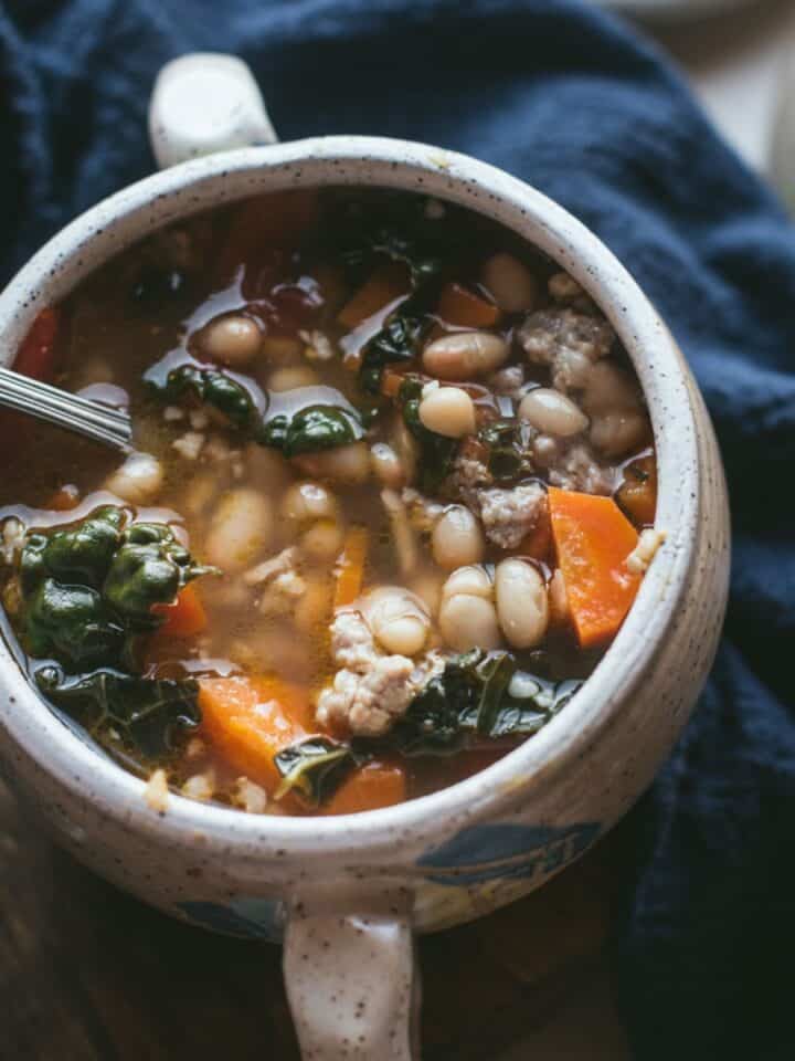 A deep white double-handled bowl is filled to the top with Sausage, White Bean, and Kale Soup. You can see all the different ingredients through the broth liquid. The bowl is on a wooden counter and a blue table cloth is laying next to it.