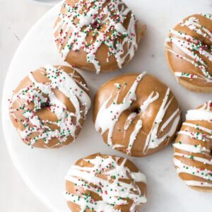 View form above of fluffy baked gingerbread doughnuts. 5 doughnuts in a ring with a 6th in the middle on a large white plate. Each doughnut is topped with a brown colored gingerbread glaze and white icing has been drizzled decoratively on top, along with festive colored sprinkles. To the top left of the doughnuts is a white bowl filled with doughnut holes.