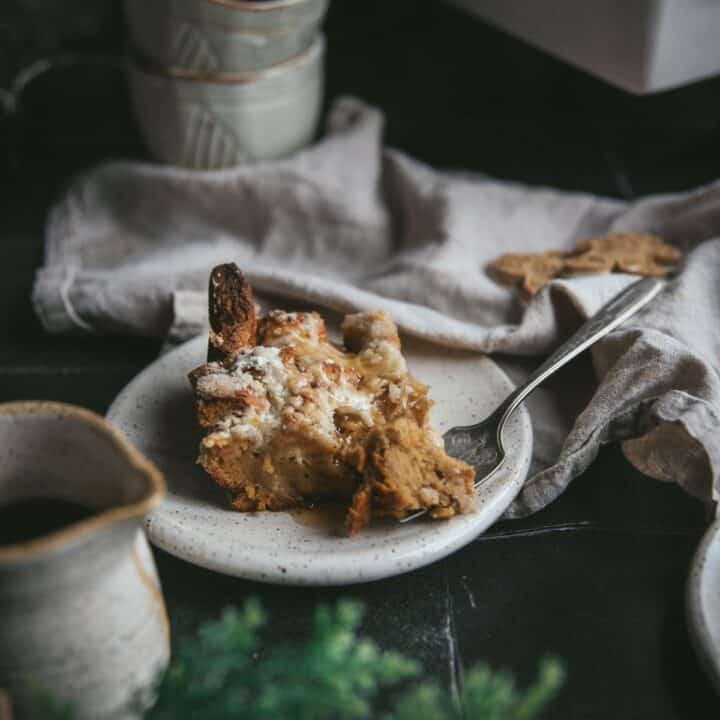 A piece of Gingerbread French Toast Casserole on a white ceramic plate. An antique fork has a piece of the french toast on it. The plate is on green tile surrounded by linen napkin, cups and gingerbread cookies.
