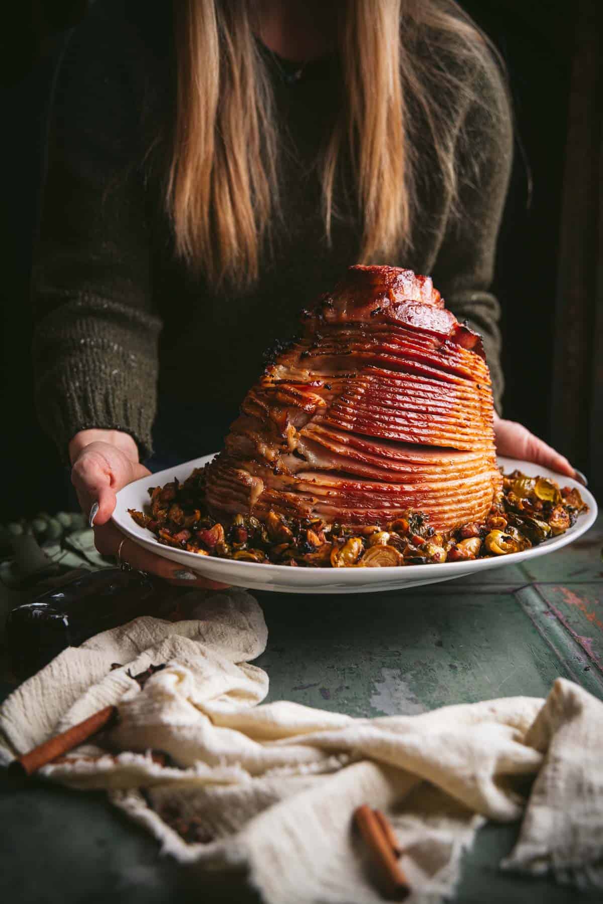 A lady is holding the bourbon smoked ham placed vertically on a white plate, she is putting the plate on the table. The ham is sitting on a bed of crispy brussels sprouts.