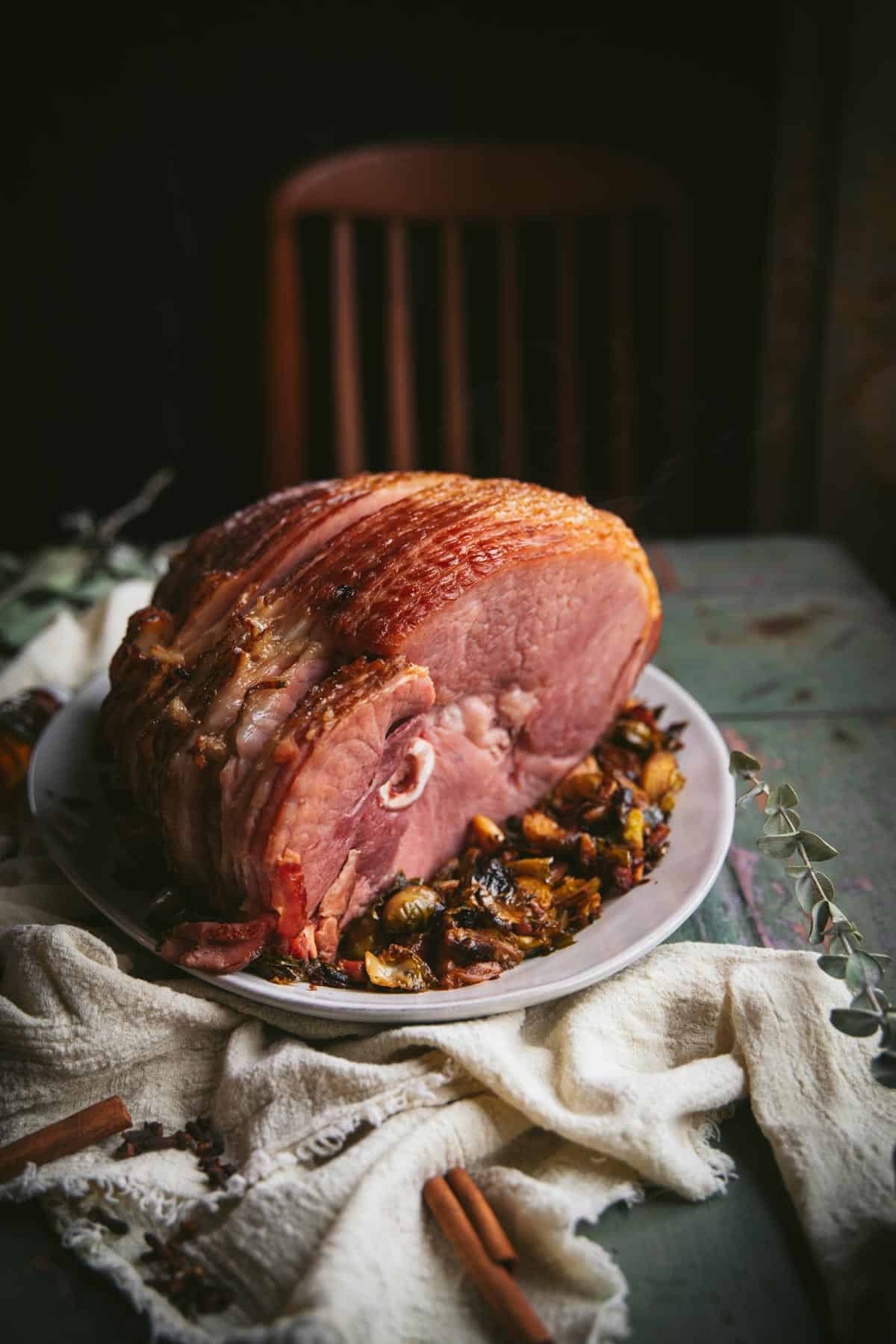 The finished brown sugar bourbon glazed ham is sitting on the table, on a white plate, on a bed of crispy sprouts.