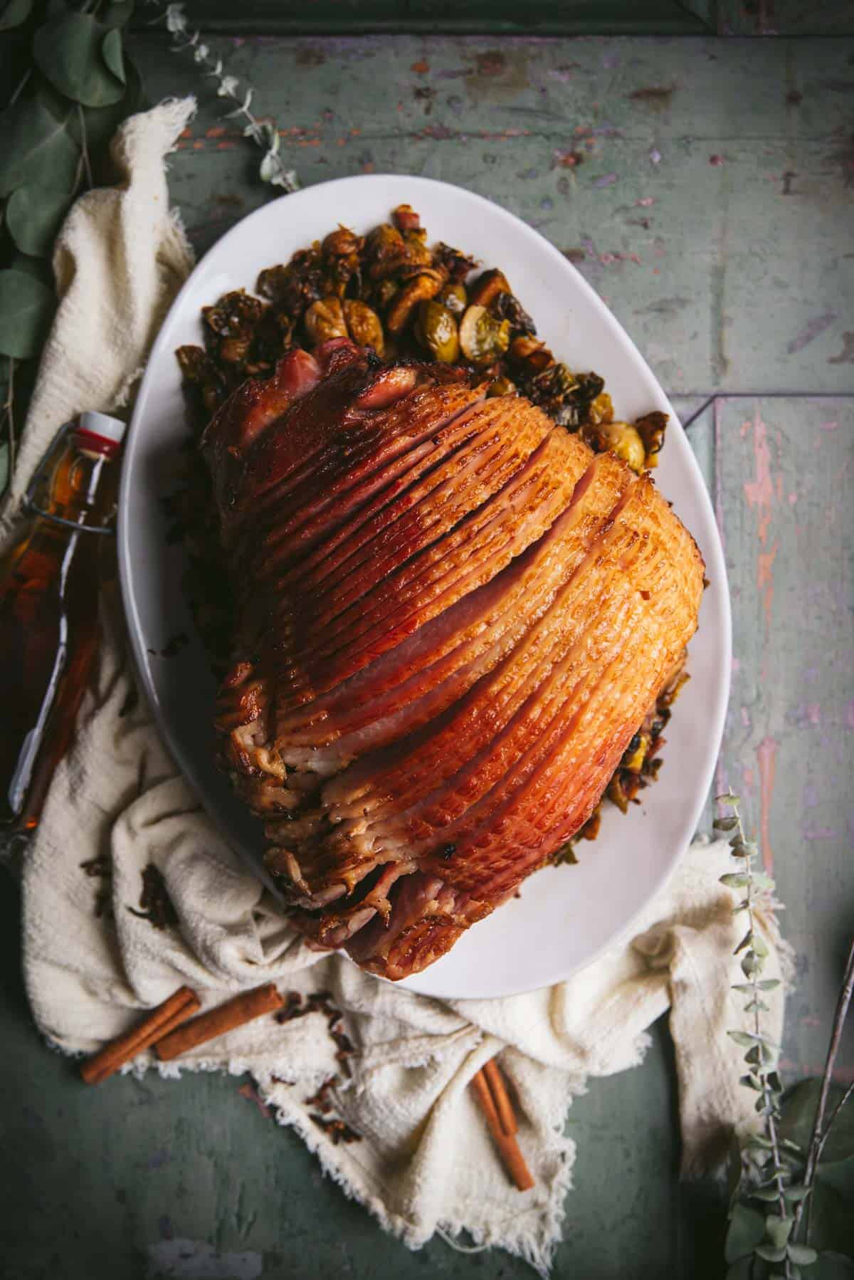 Ariel shot of the brown sugar and bourbon glazed ham. It is sliced into thin pieces and sitting on a large white plate. Next to the plate is a brown glass bottle and white cloth.