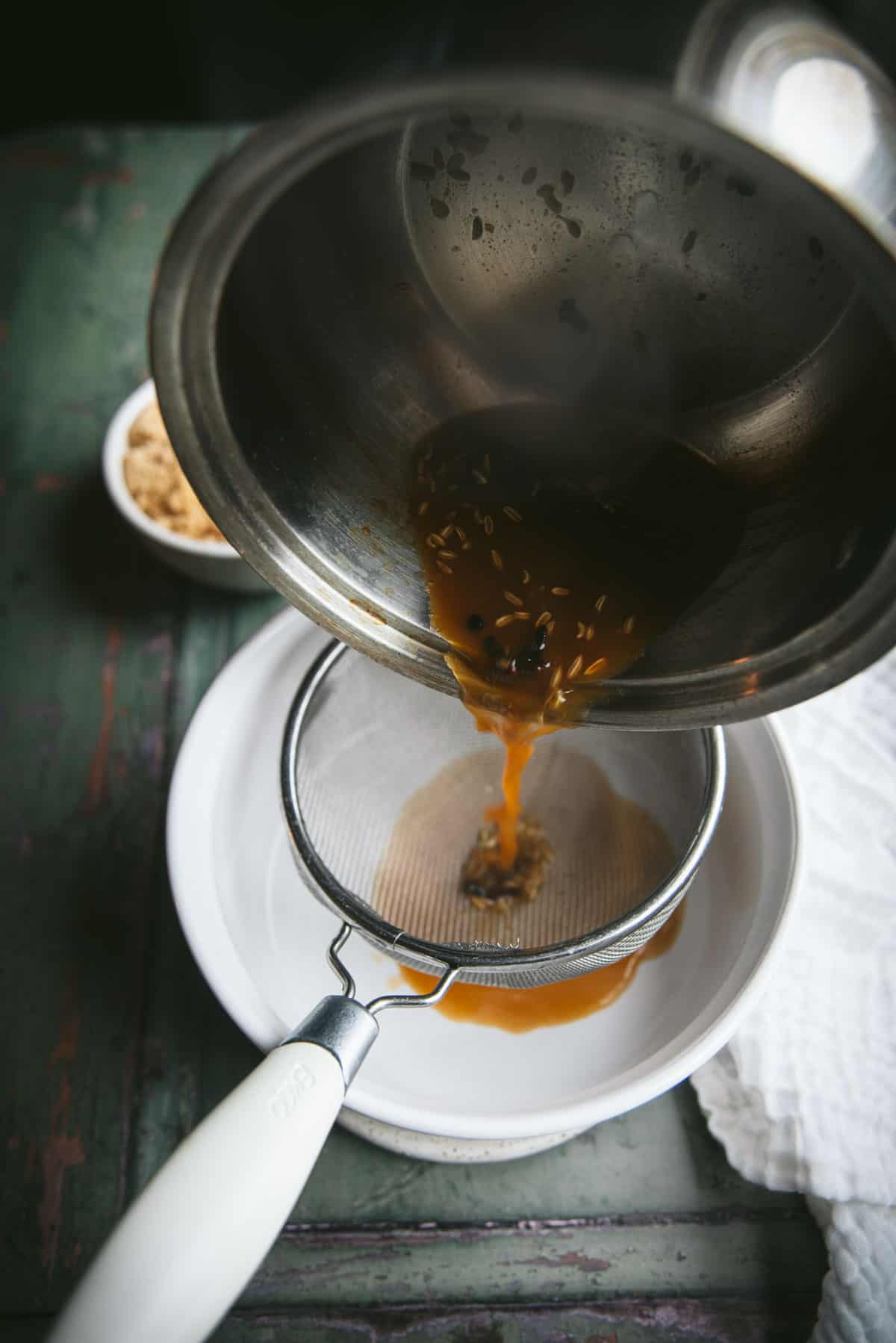 The liquid spice mixture is being poured from the silver pot, through a metal strainer and the liquid is being collected in a white ceramic pot underneath.