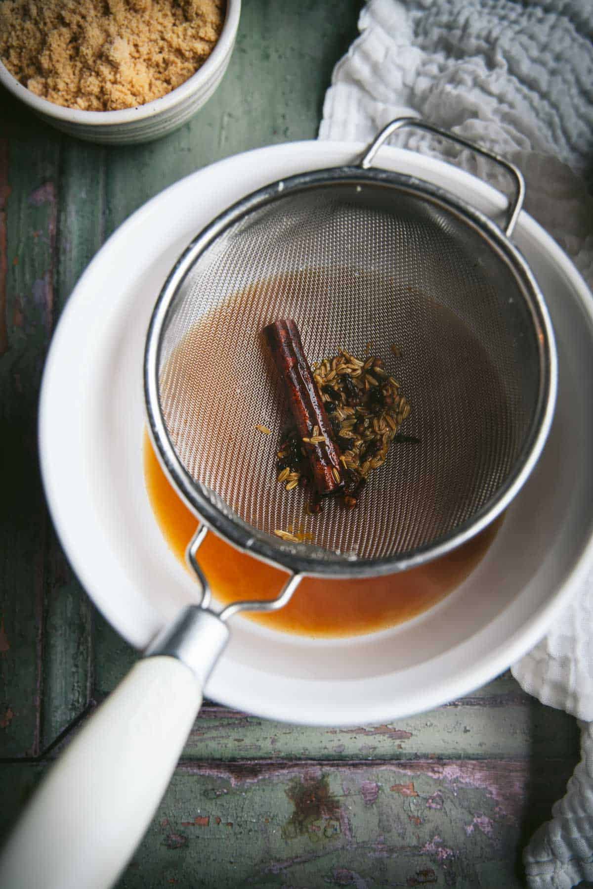 There is a silver metal strainer sitting on top of a white ceramic bowl. In the bottom of the bowl is amber colored liquid. The spices that were strained out of this liquid are sitting in the strainer in top.