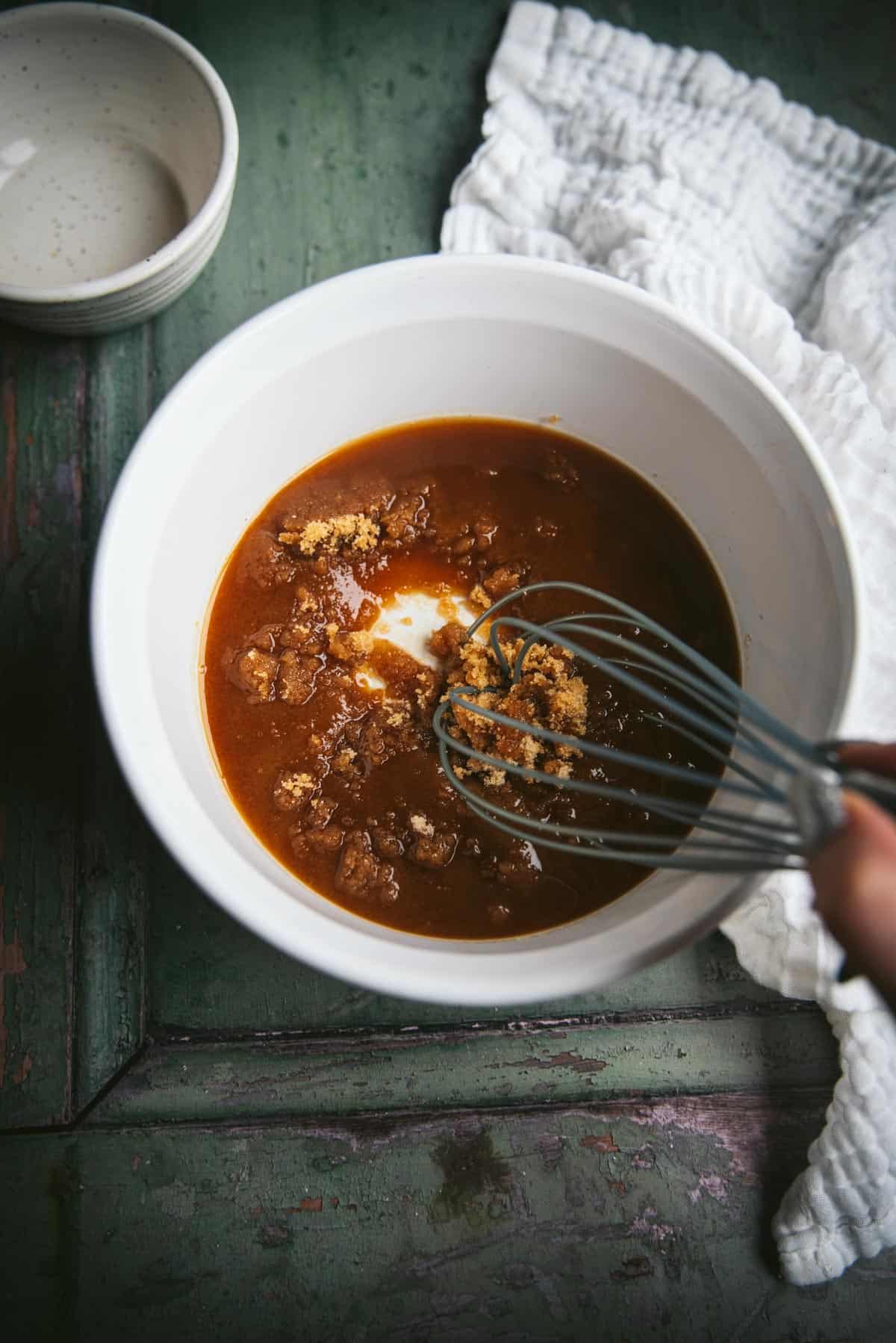 Someone is whisking brown sugar and the spiced liquid together in a large white ceramic pot.