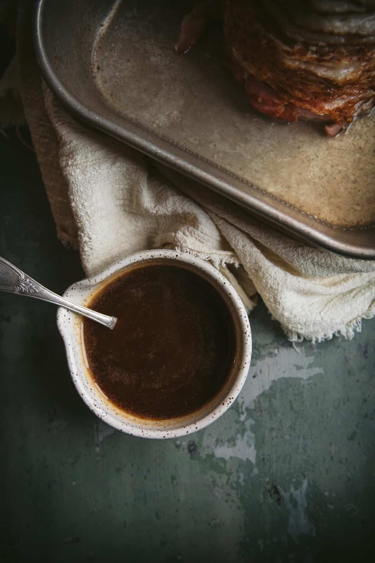 The glaze is sitting in a white ceramic dish with a spout. There is a silver spoon resting in the bowl too. In the background you can see the corner of the baking tray where the ham is.