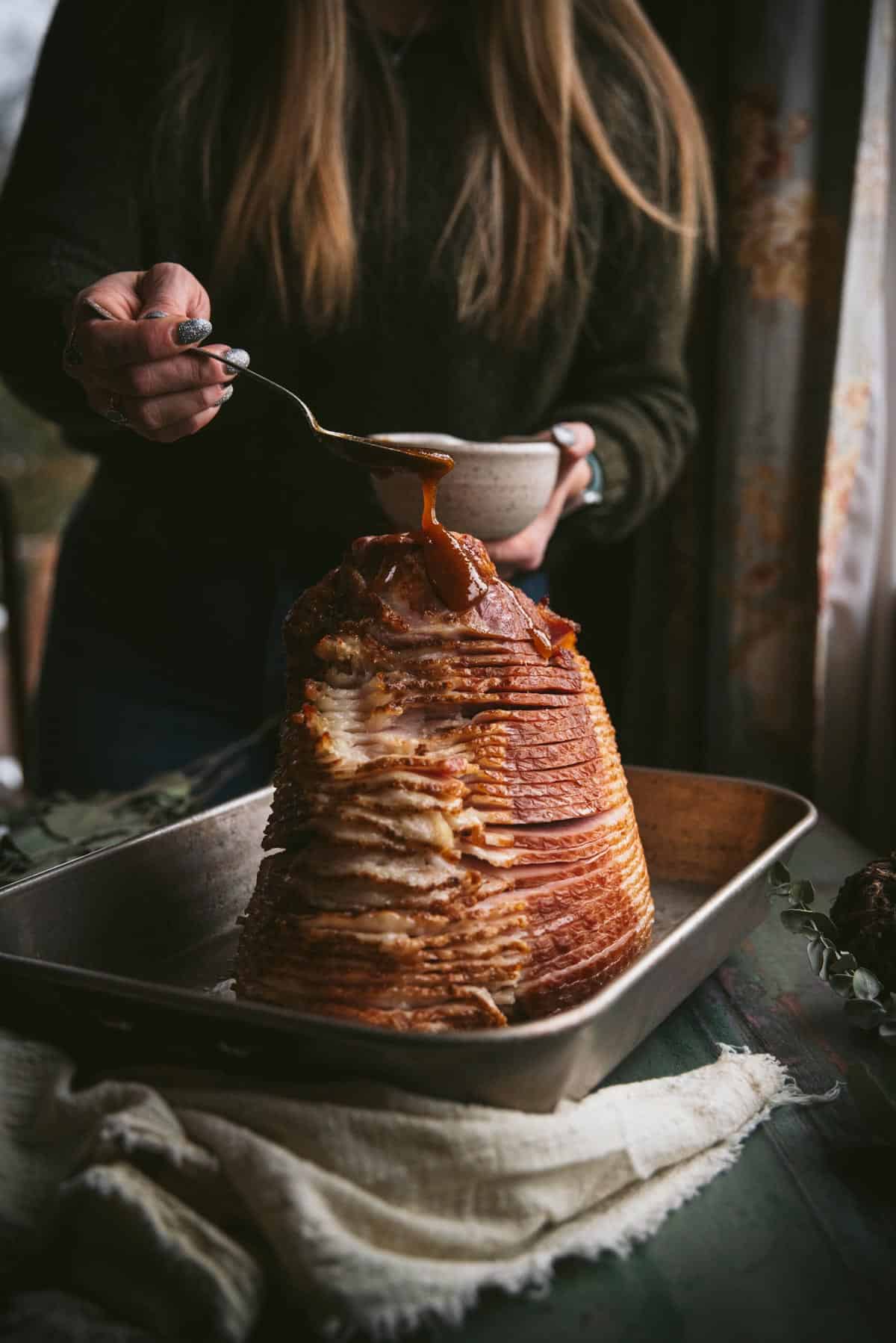 The ham is standing upright in a baking tray and is sliced partially. A woman in a green jumper is pouring the bourbon glaze over the top with a spoon.