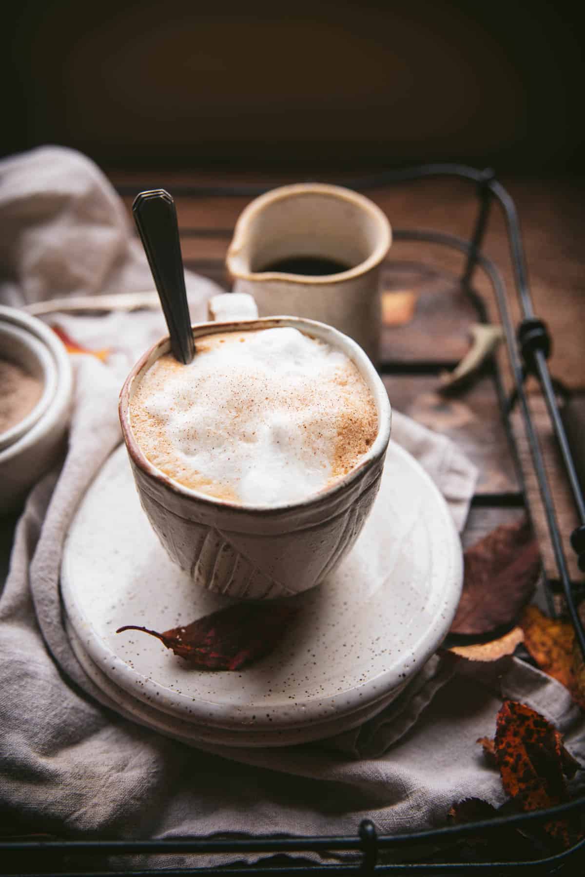 Milk foam sprinkled with maple sugar in a white ceramic mug on top of a white ceramic plate.  There's a spoons sticking out of the much and the mug and other plates and dishes are on a wood and wire service tray.