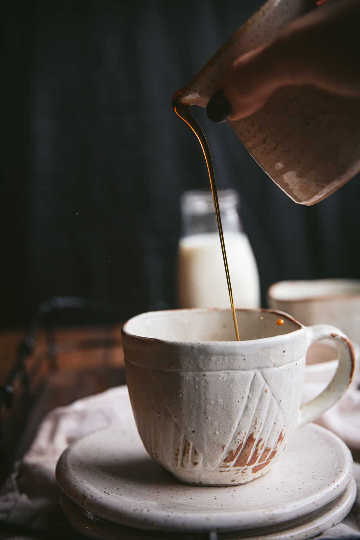 Cermaic carafe pouring maple syrup into a ceramic mug.