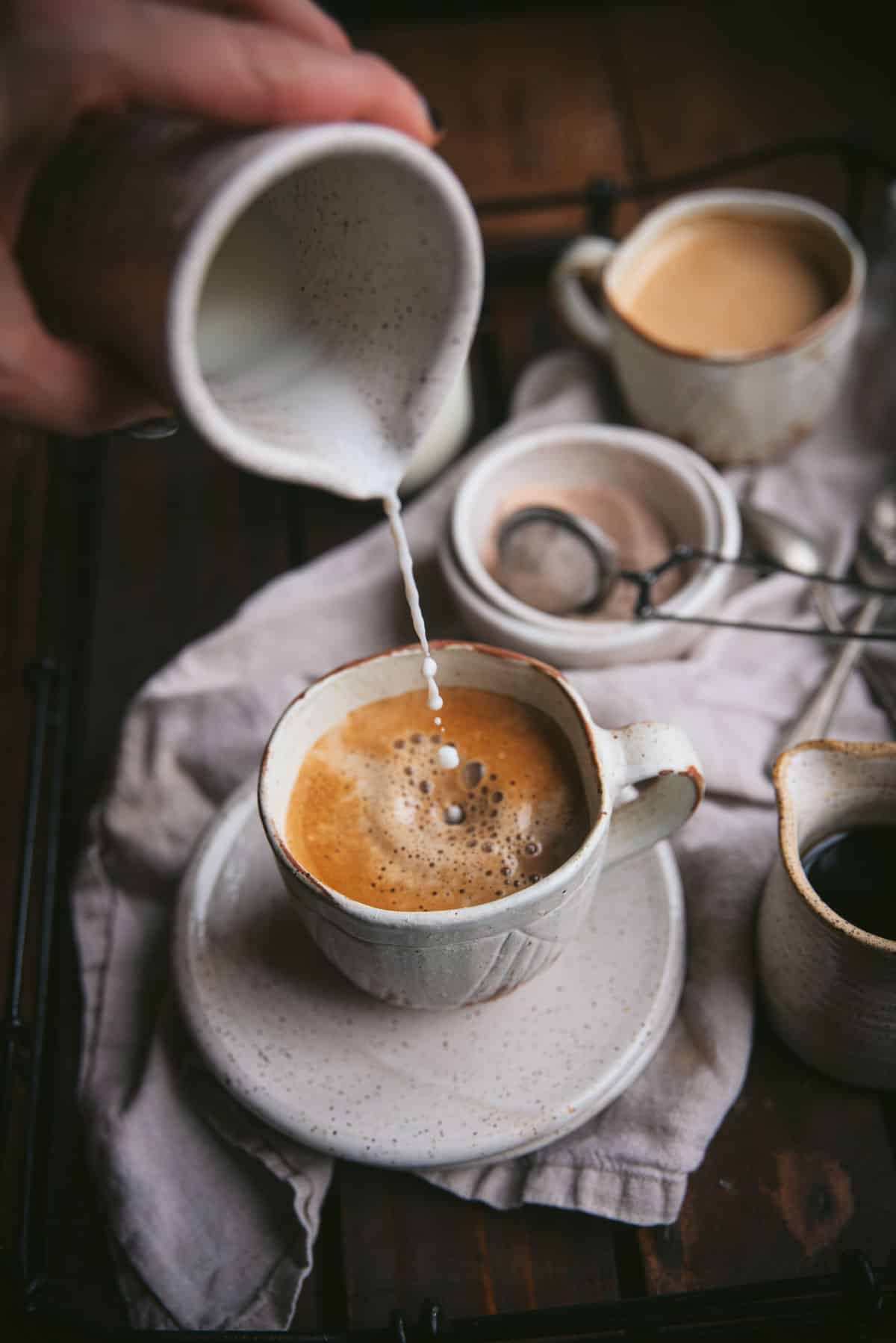 Hand held ceramic carafe pouring steamed milk into a coffee mug with espresso and maple syrup.  The milk is being slowly poured and single drops are hitting the surface of the coffee.