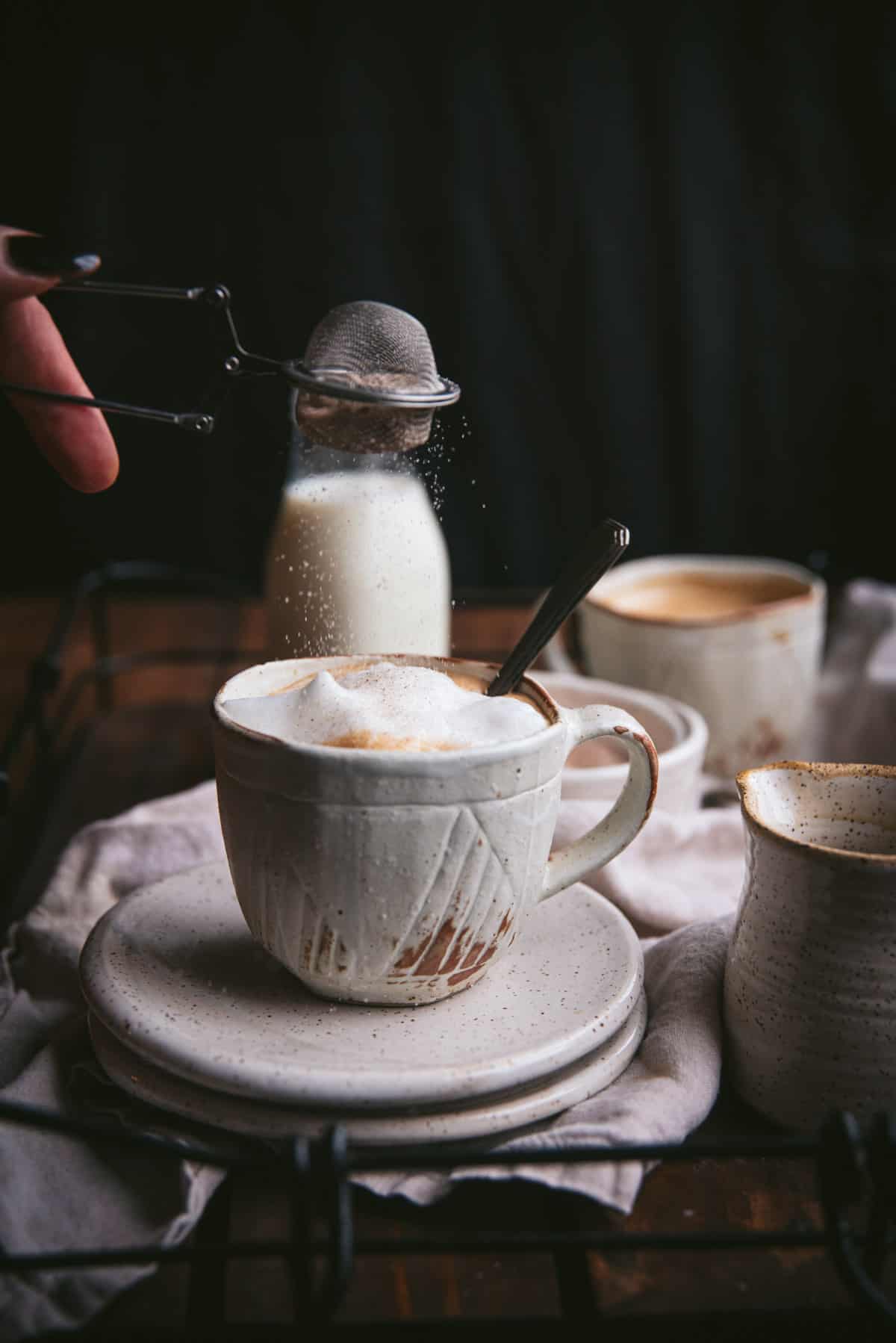 Tea strainer sprinkling maple sugar over frothy milk on top of a maple latte.  The mug is stacked on two ceramic plates.