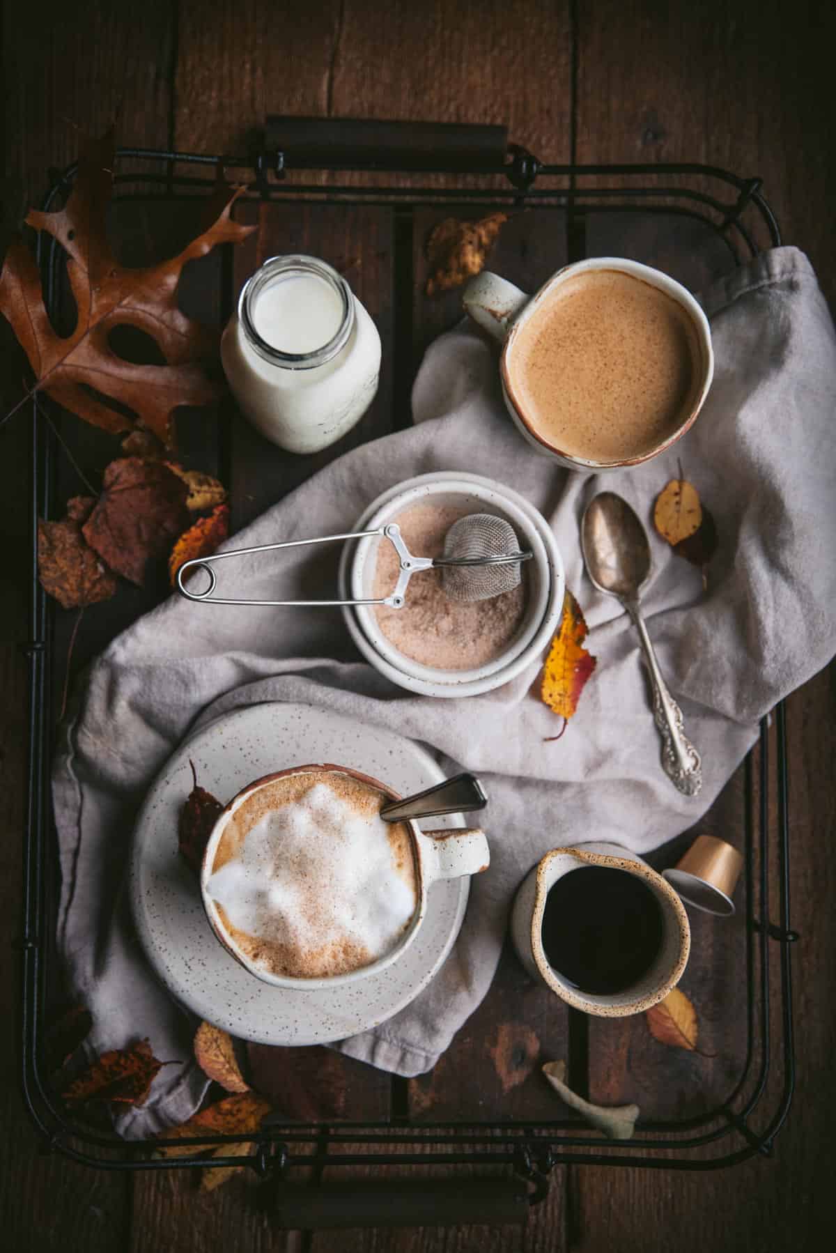 Overhead image of a wooden and wire tea tray topped with a tan linen napkin, maple leaves, and ceramic containers with ingredients for maple lattes.  Two coffee mugs contain lattes.