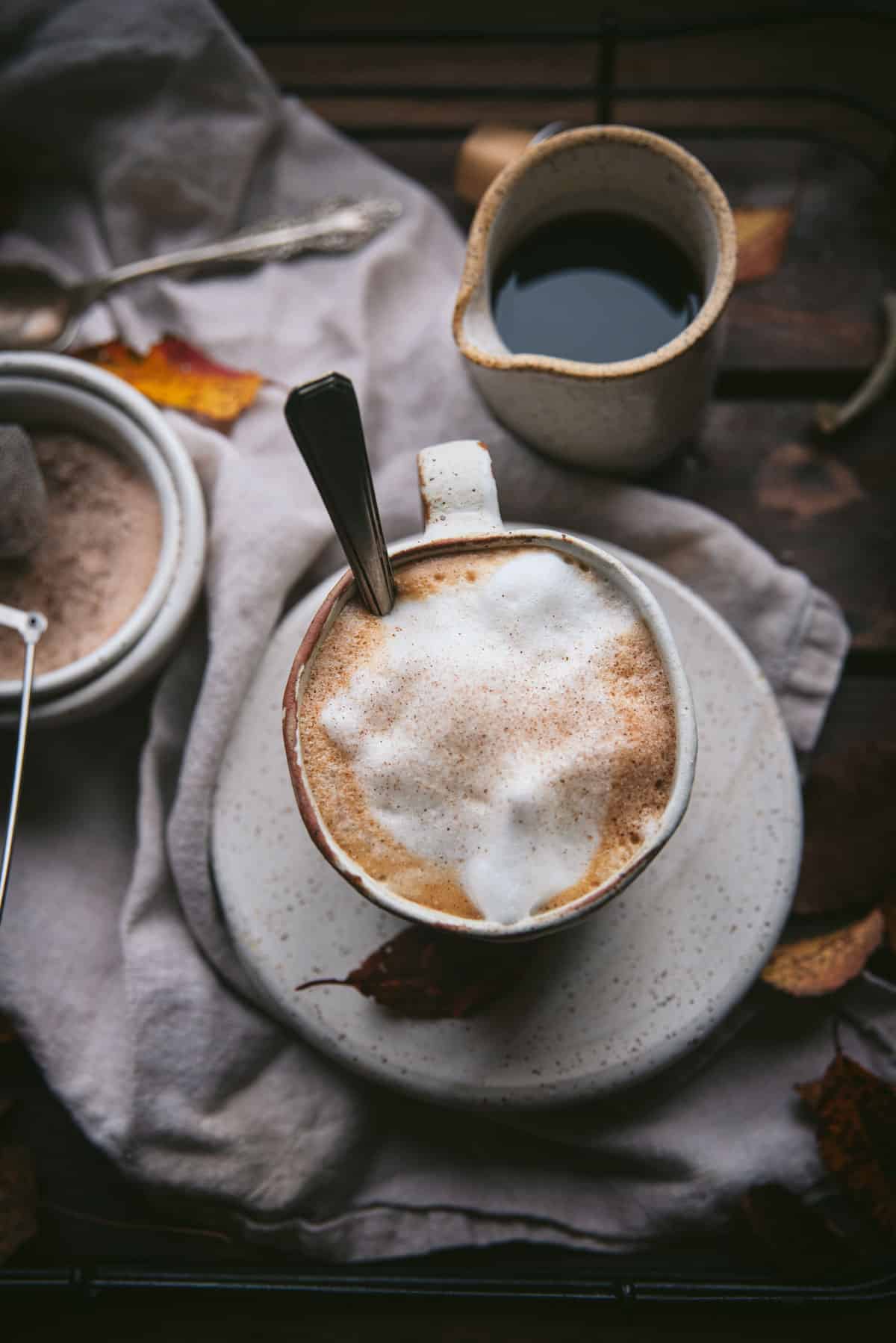 Close up overhead image of maple latte in a white coffee mug on top of stacked ceramic plates.  Maple syrup is in a cermaic pourer behind the cup.