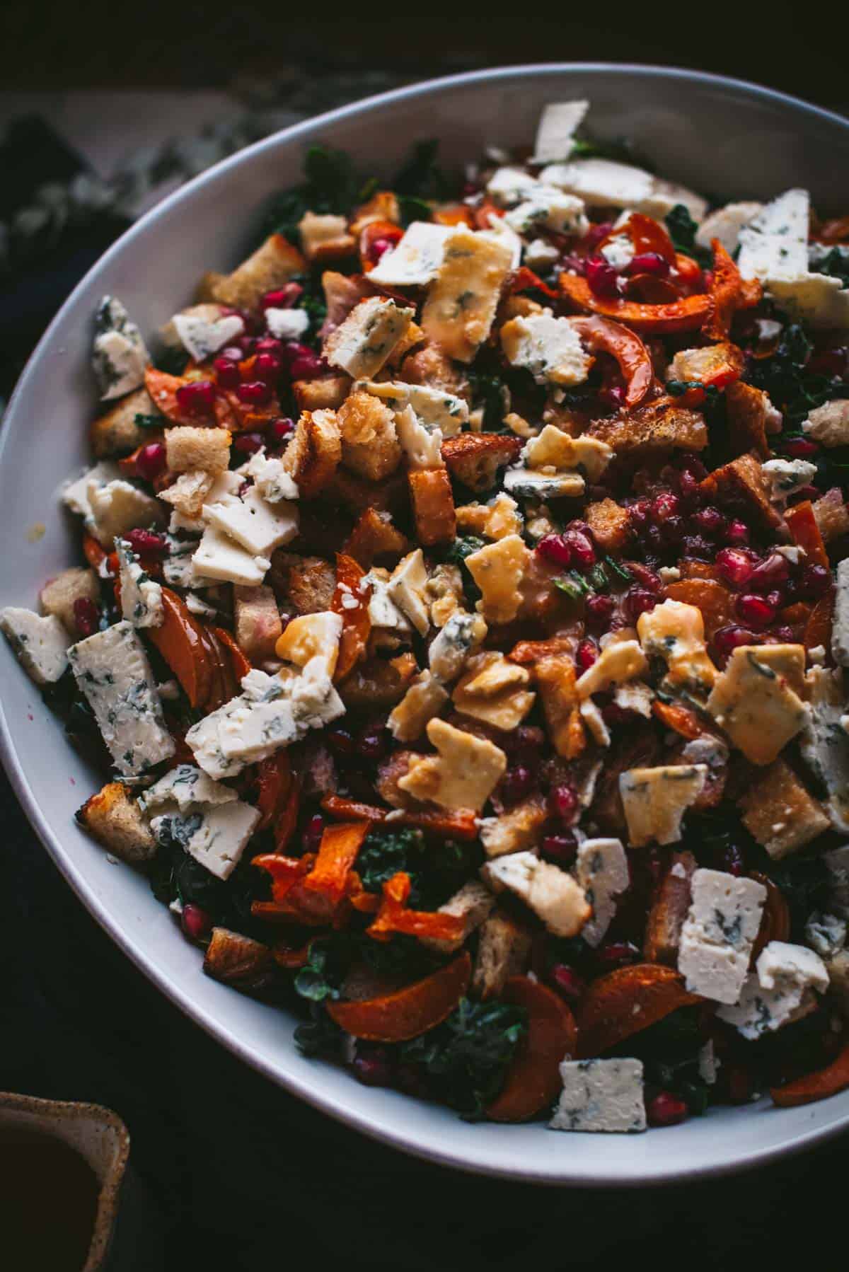 overhead image of a large bowl of salad with kale, blue cheese, croutons, pomegranate, roasted squash, and maple miso dressing poured over it.