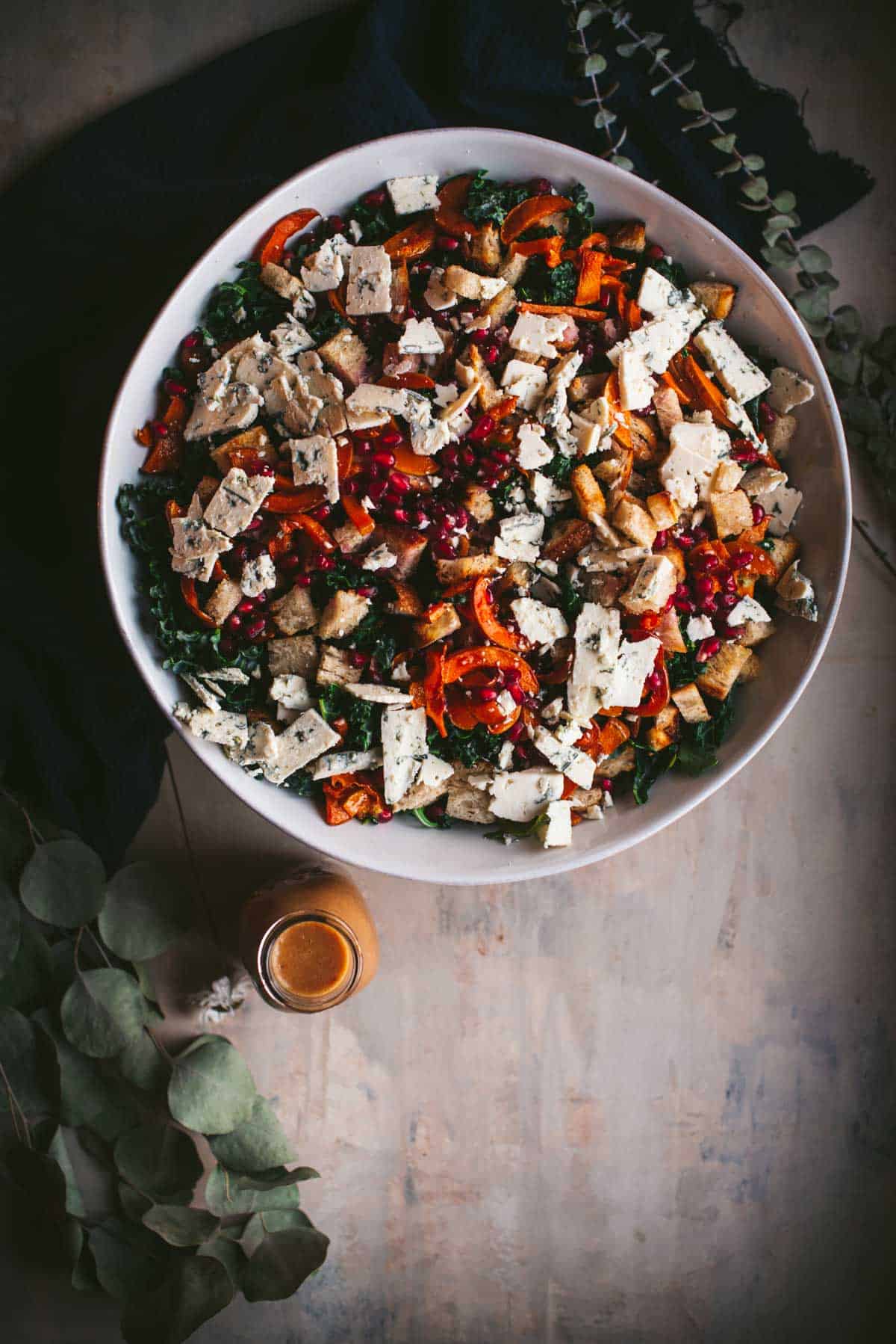 overhead image of a large bowl of salad with kale, blue cheese, croutons, pomegranate, roasted squash, and maple miso dressing in a glass container next to it.