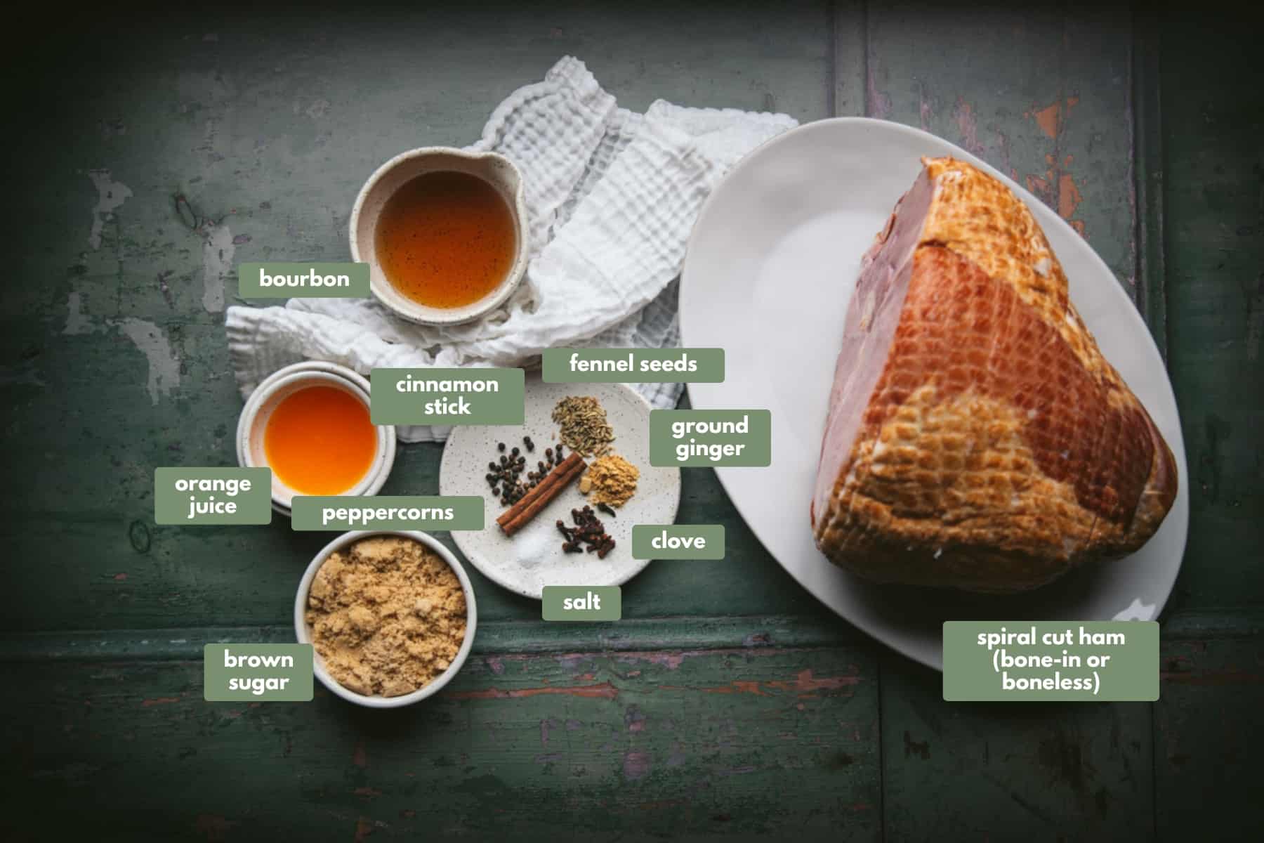 All  the ingredients required to make this glazed ham recipe. 
There is a large spiral cut ham sitting on a large white plate, a white dish filled with bourbon, a small white bowl with orange juice, a white bowl filled with brown sugar and a white plate containing spices: cinnamon sticks, fennel seeds, ground gingr, cloves and salt.