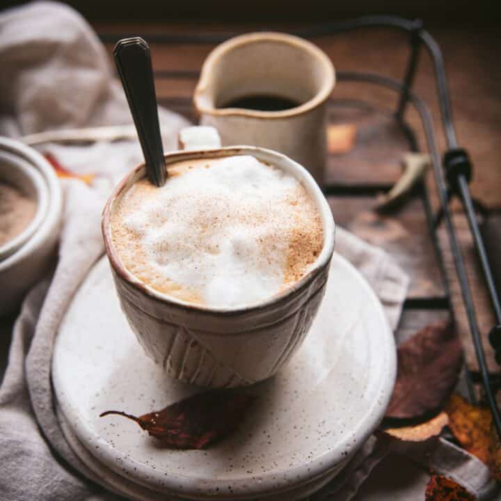 Milk foam sprinkled with maple sugar in a white ceramic mug on top of a white ceramic plate. There's a spoons sticking out of the much and the mug and other plates and dishes are on a wood and wire service tray.