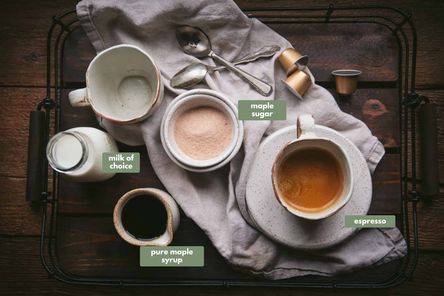 Ingredients for maple lattes in ceramic bowls, mugs, and containers on top of a tan linen napkin and on a wooden serving tray.