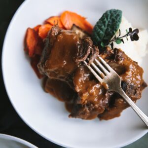 Plated beer braised short ribs on a white plate. An antique looking for has a piece on short rib with sauce on it. Underneath the fork the meat is plated with carrots and mashed potatoes.