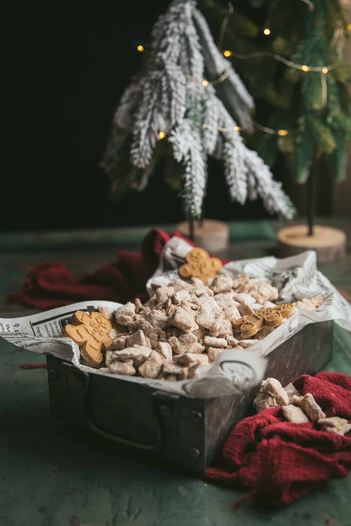 Side angle image of gingerbread puppy chow in a parchment lined tin container.  There are mini christmas trees with lights on them in the background.