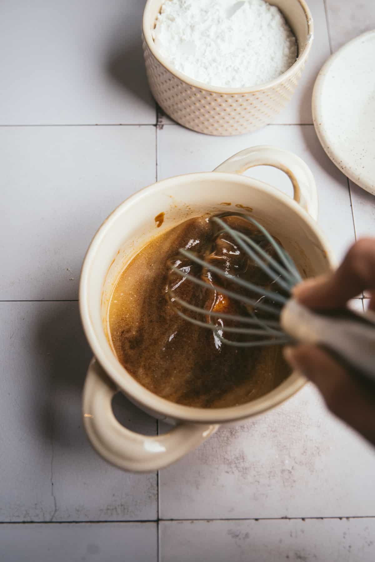 a hand using a whisk to mix melted butter and molasses in an off white bowl with two handles.  a container of powdered sugar and a plate are in the background.