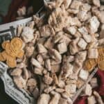 verhead image of Chex cereal coated with white chocolate, spices, and powdered sugar, with an antique serving spoon and gingerbread shaped cookies on top of parchment paper in a serving dish
