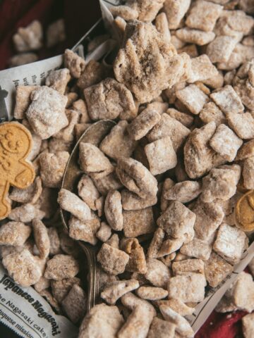 verhead image of Chex cereal coated with white chocolate, spices, and powdered sugar, with an antique serving spoon and gingerbread shaped cookies on top of parchment paper in a serving dish